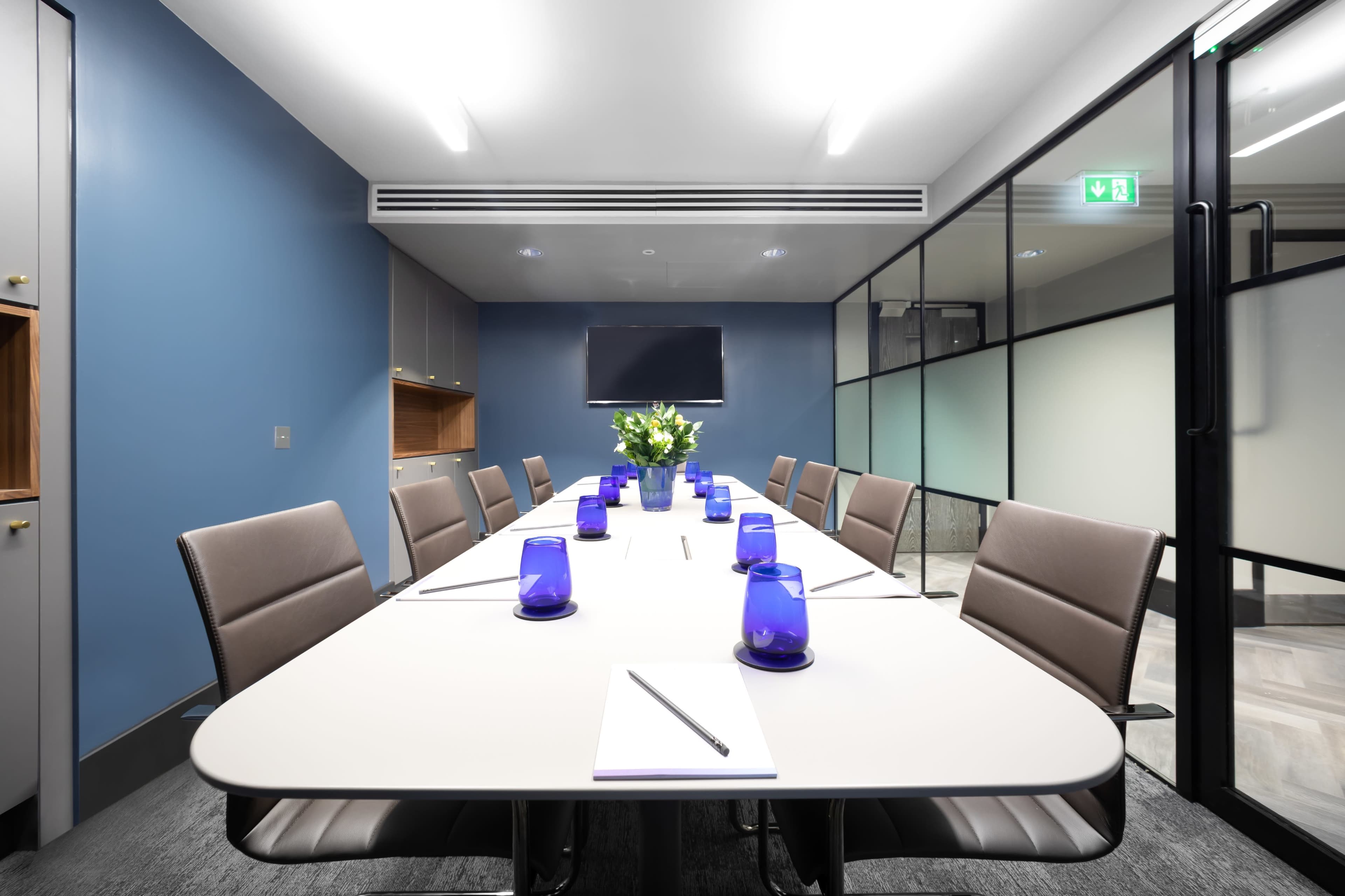 A modern conference room features a long table set with blue candle holders and notepads, surrounded by sleek chairs and a wall-mounted television.