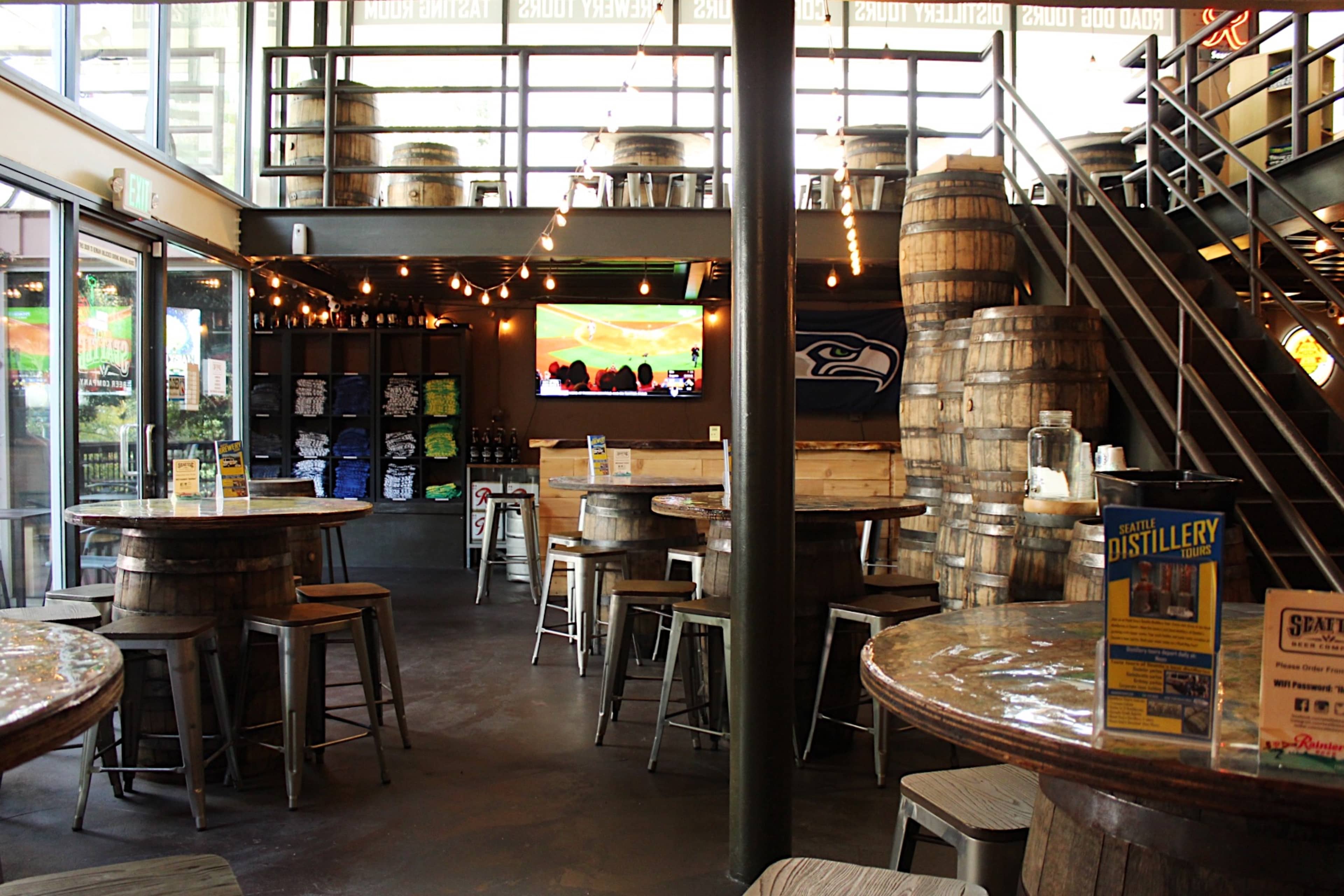 The interior of a brewery features wooden barrel tables and stacks of barrels, with a television displaying a sports event and merchandise on display.