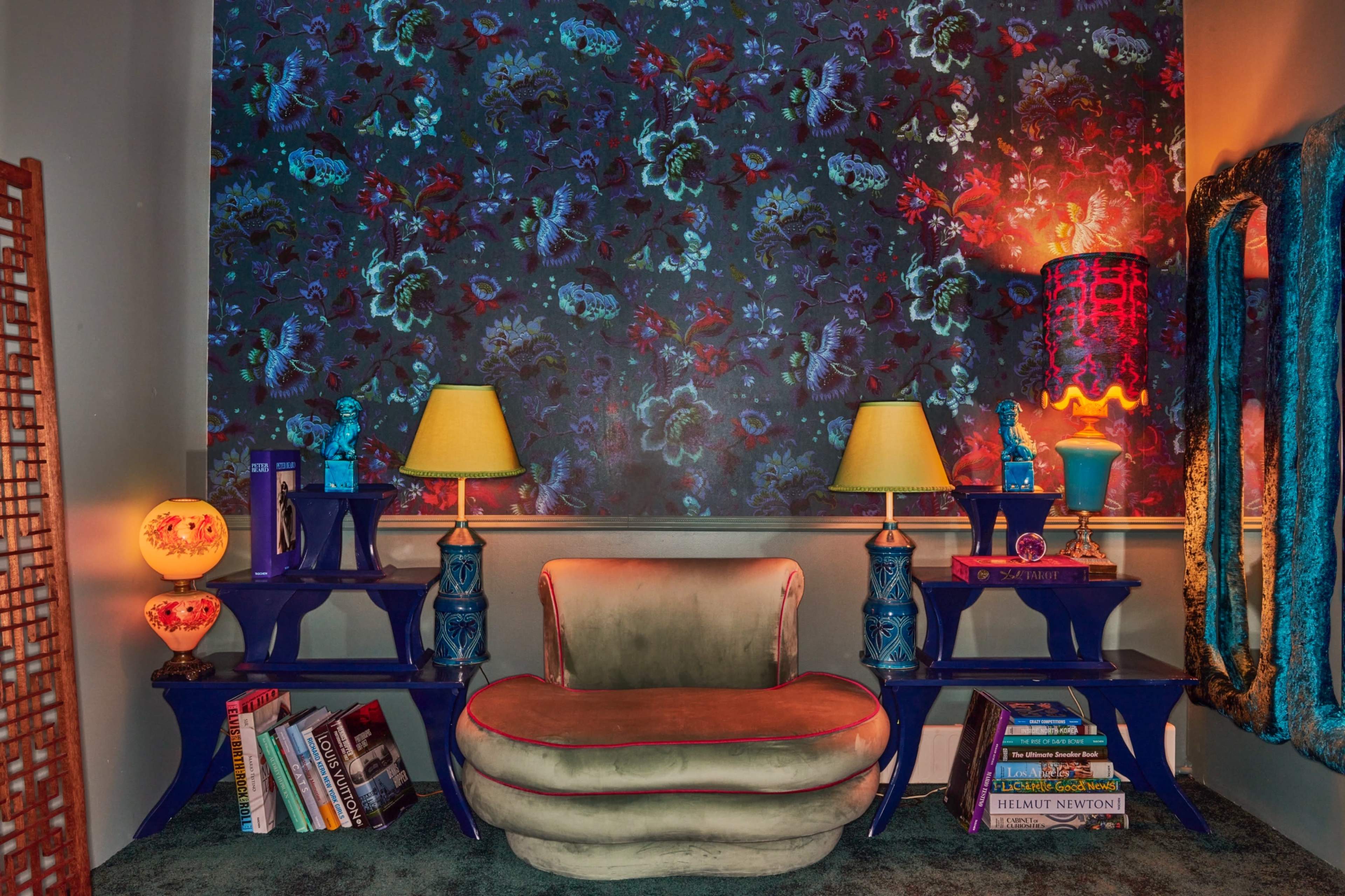 A stylish seating area with a curved chair, colorful lamps, and decorative shelves adorned with books and accessories against a vibrant floral wallpaper.