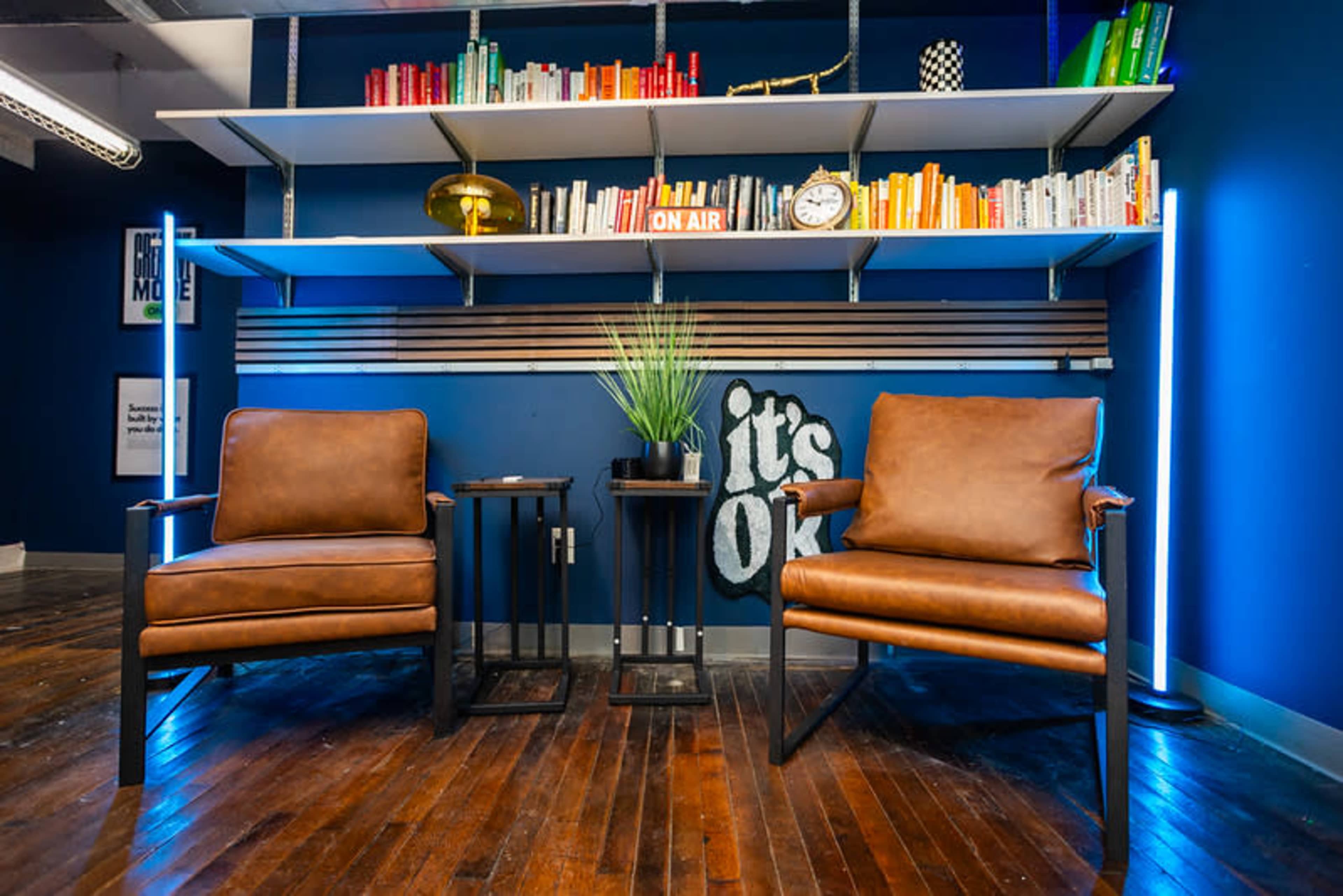 The image shows a cozy interior space featuring two brown leather chairs, a small side table, and a bookshelf filled with colorful books against a blue wall.