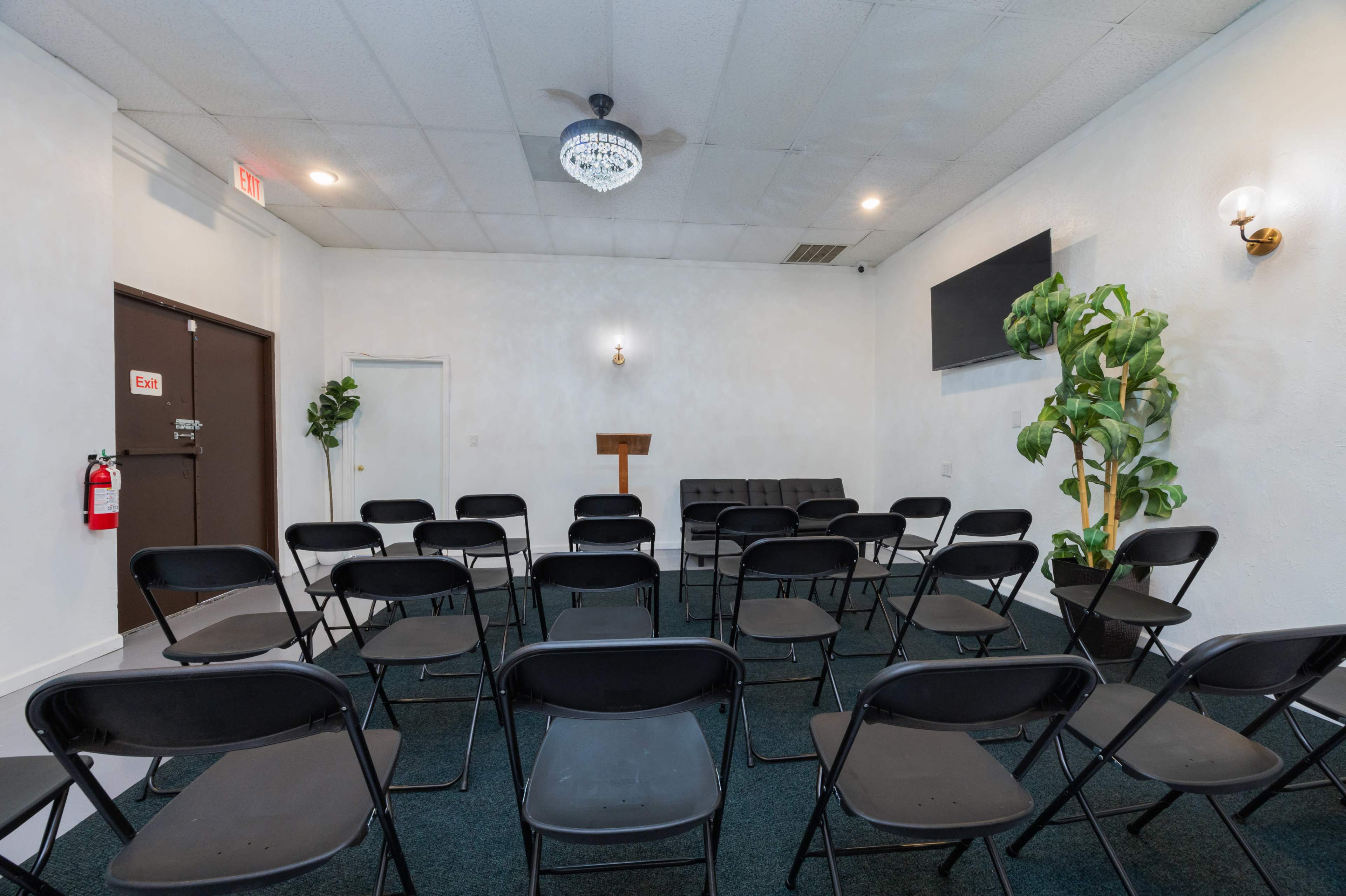 A small, empty meeting room arranged with black folding chairs facing a wooden lectern, surrounded by a plant and featuring a wall-mounted screen.