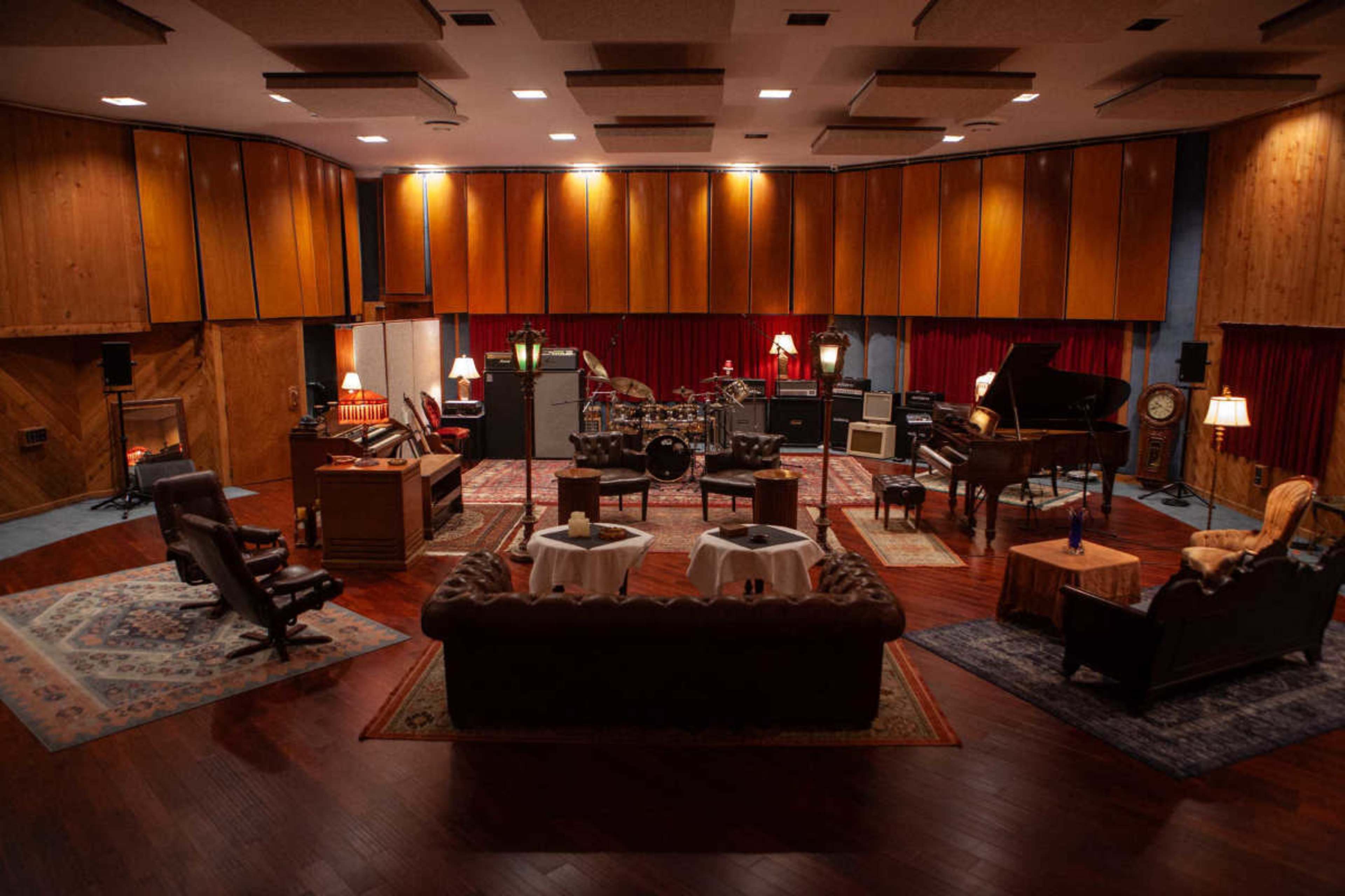 The image shows a spacious music recording studio with wooden paneling, vintage furniture, and various musical instruments arranged in the center.