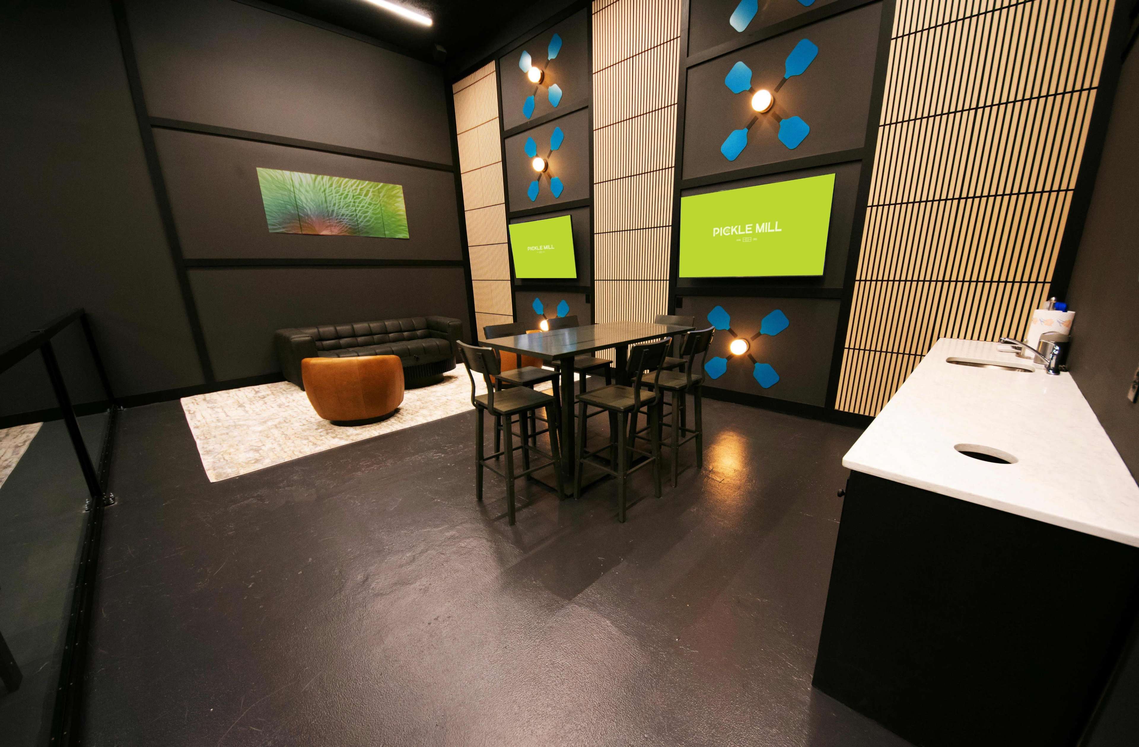 The image shows a modern meeting room featuring a black sofa, a round pouf, a large table surrounded by high chairs, and two screens displaying a logo, all set against a dark-walled backdrop with decorative wall panels.