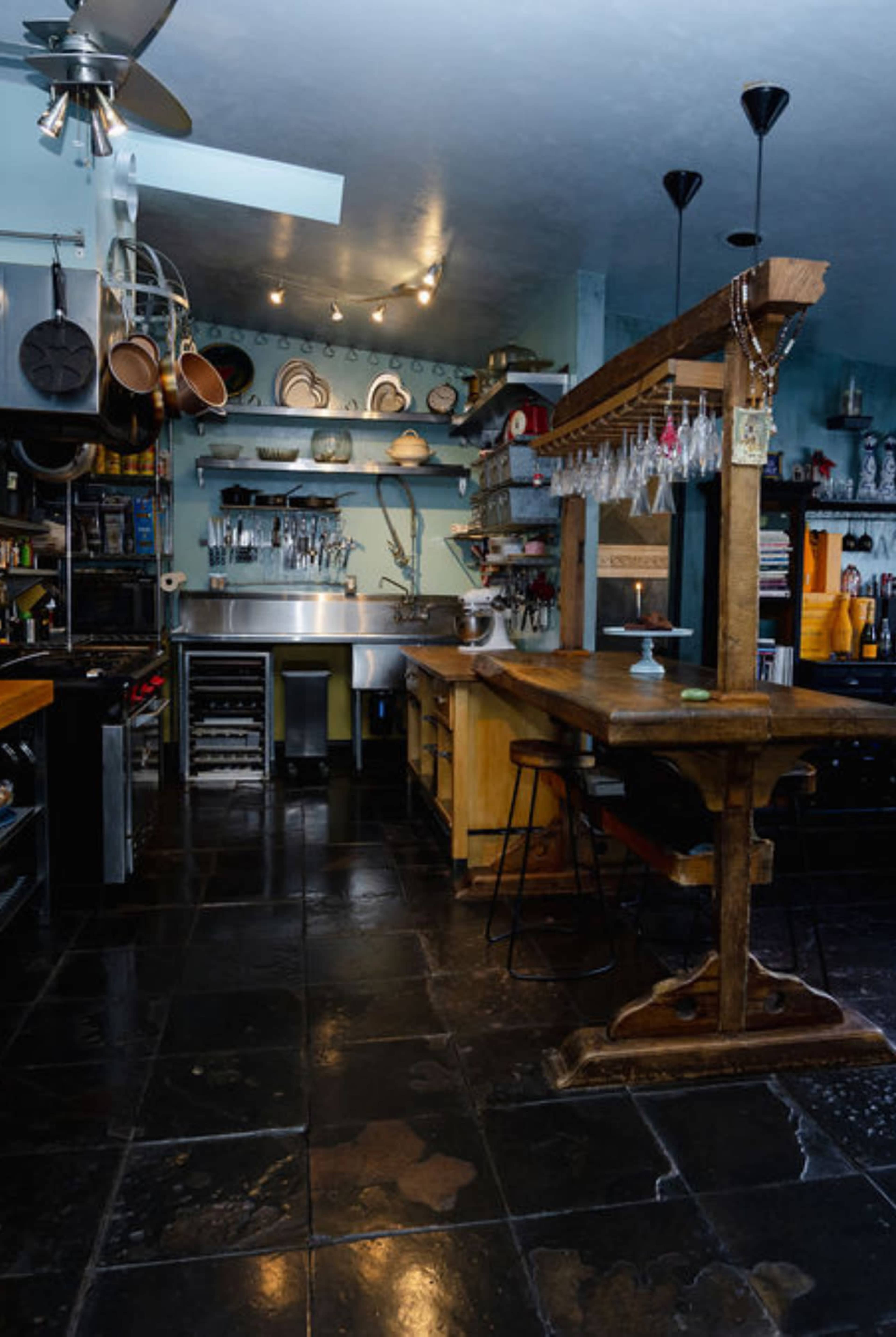 The image shows a kitchen with a large wooden table, metal shelves filled with utensils, and a stainless steel counter area.