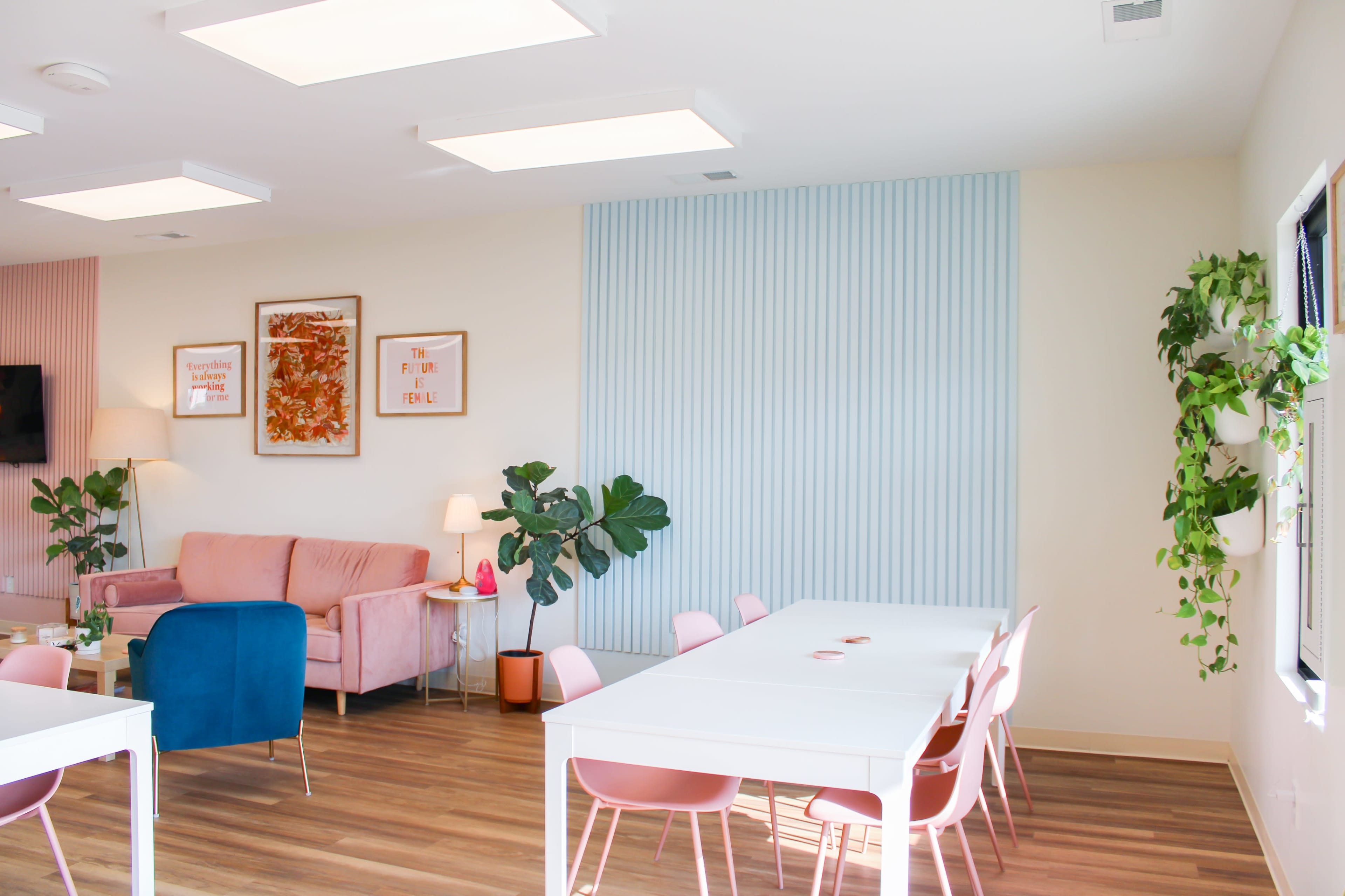 A modern interior space featuring a pink sofa, white tables and chairs, potted plants, and decorative wall elements.