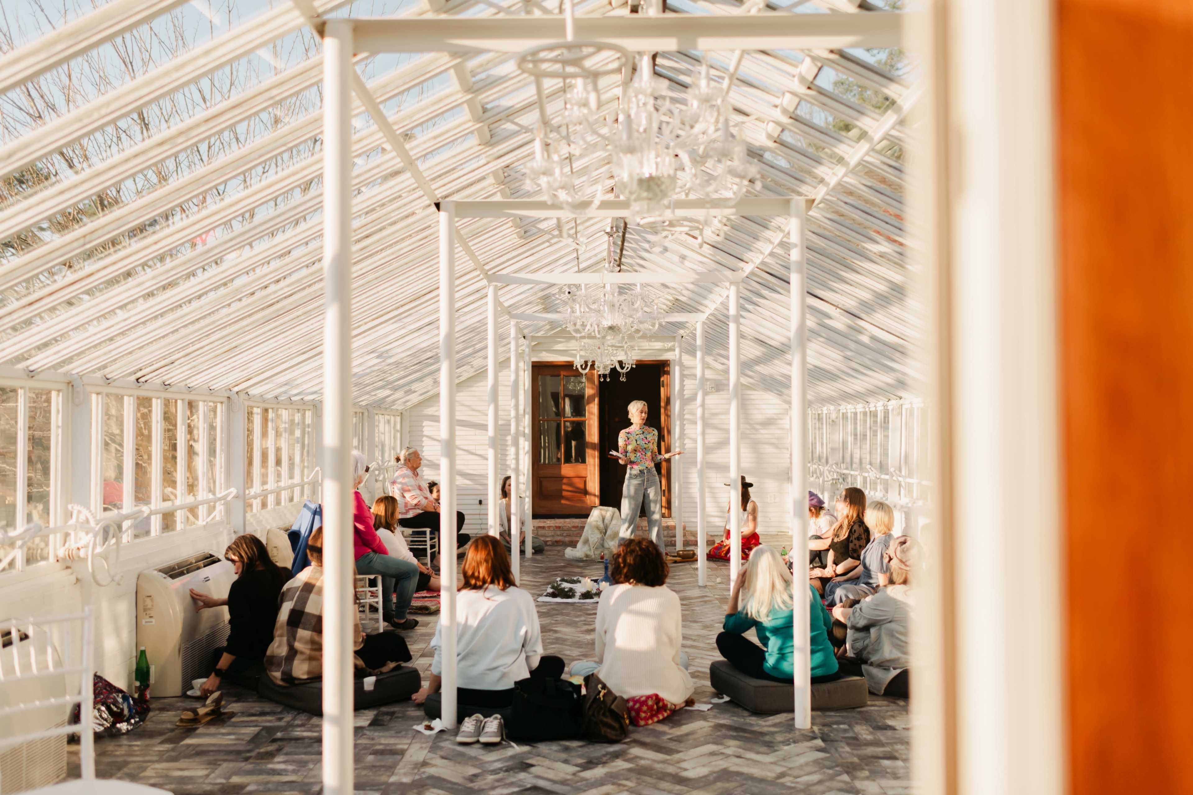 A group of people sits on the floor in a bright glass greenhouse while someone stands at the front, holding a bouquet of flowers.