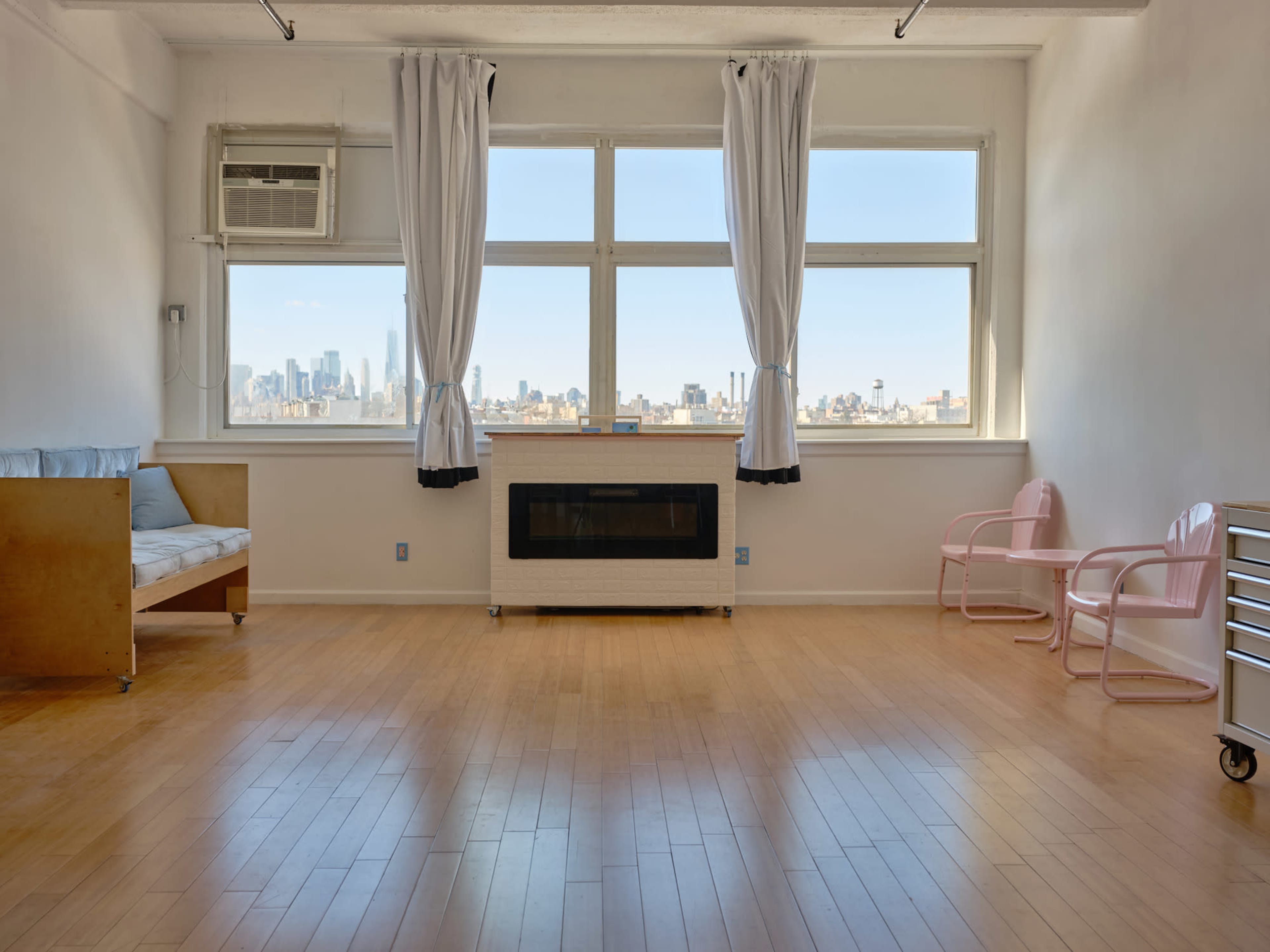 The room features large windows with city skyline views, a wooden seating area, a pink chair, and a metal storage cart.