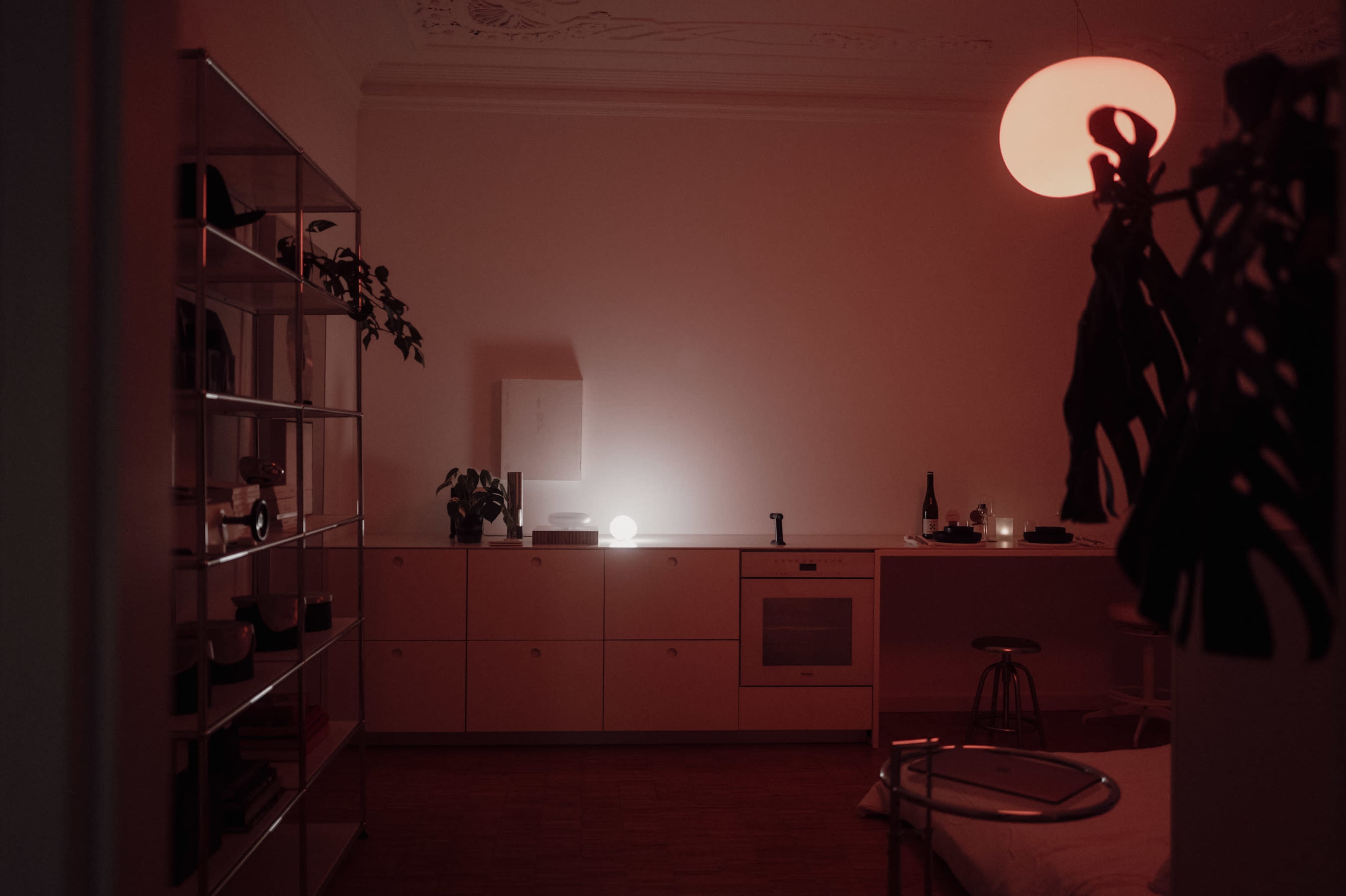 The image shows a dimly lit room featuring a modern kitchen area, a minimalist shelving unit, and a round pendant light.