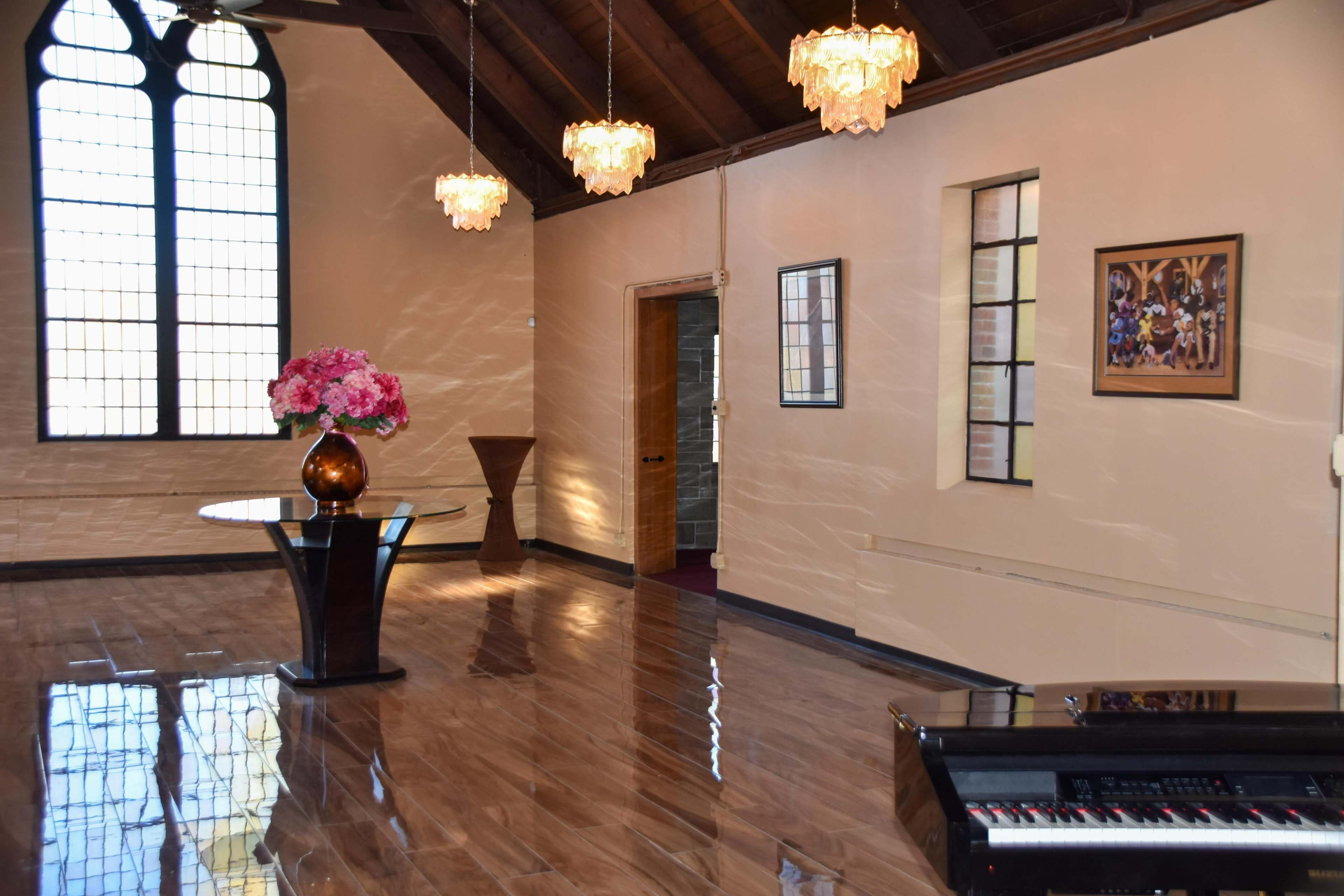 The interior features a spacious room with a high ceiling, wooden beams, large windows, a round table with flowers, and a piano in the corner.