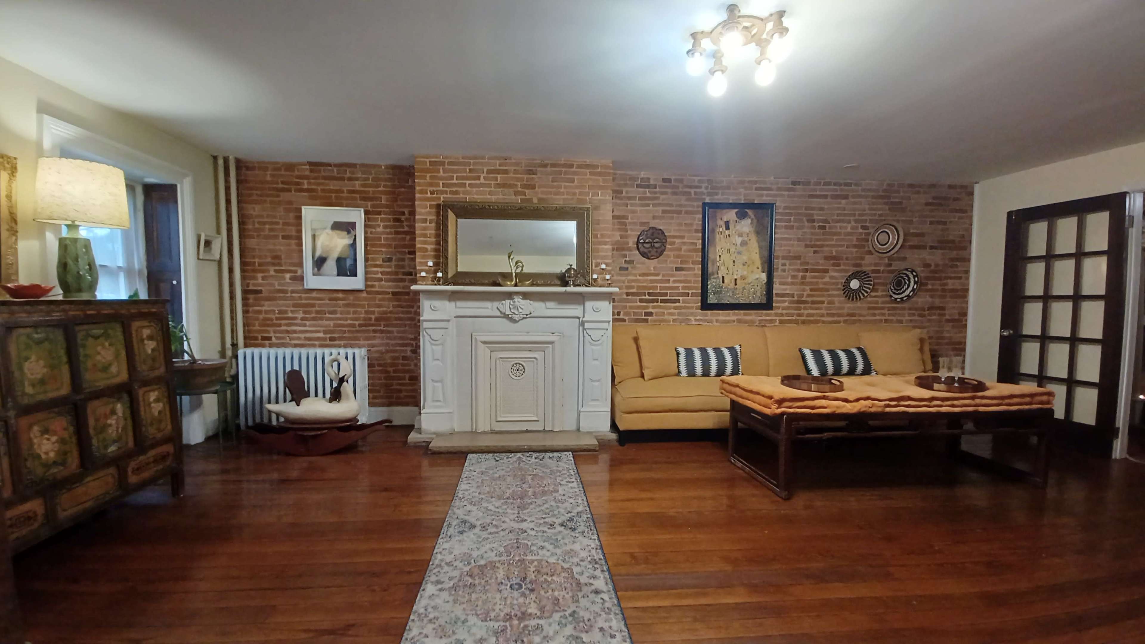 A living room with brick walls, a vintage fireplace, a yellow couch, and wooden flooring, adorned with a patterned rug and decorative artwork.