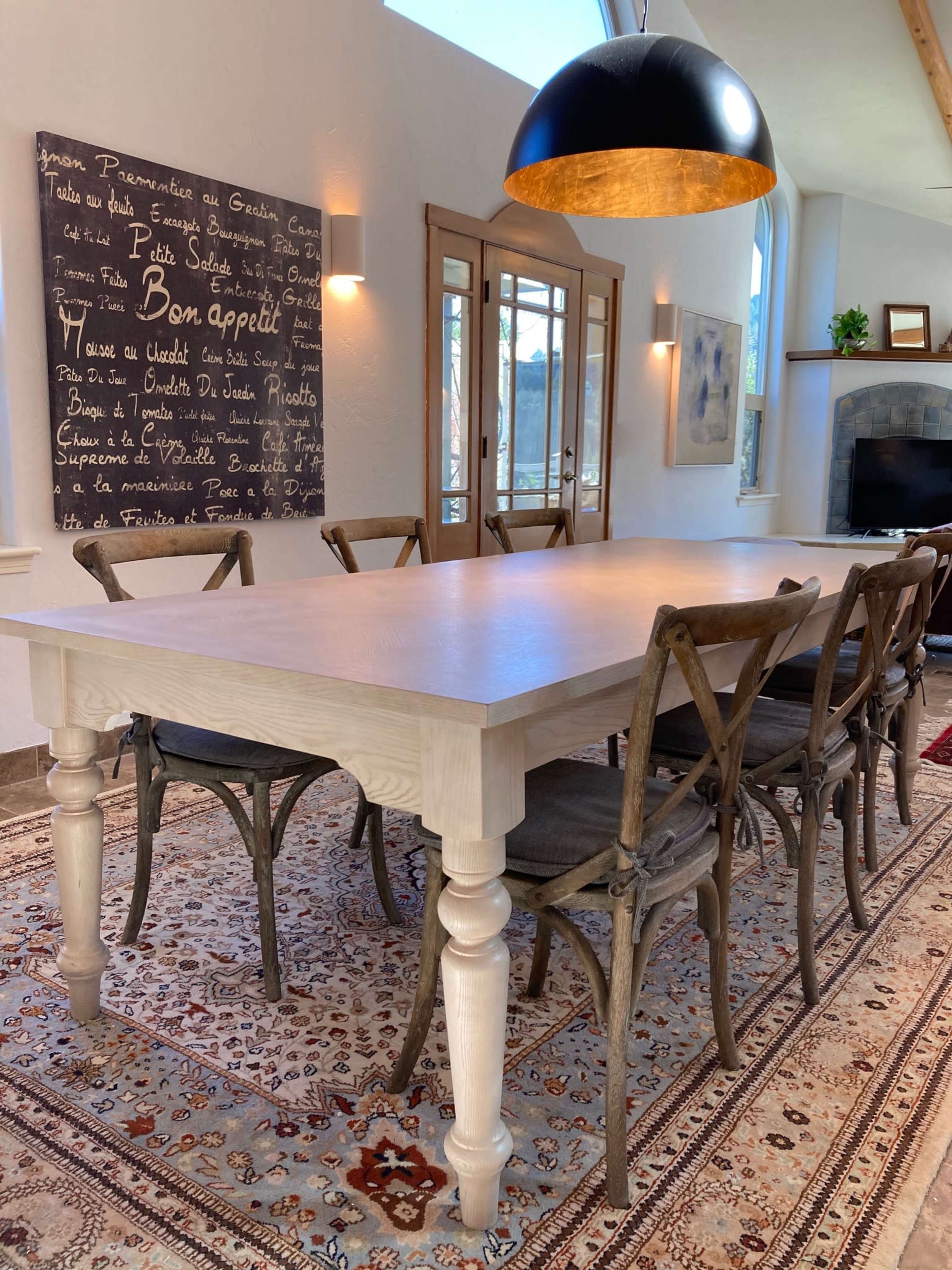 A wooden dining table with legs sits on a patterned rug, surrounded by chairs under a pendant light in a brightly lit room with large windows.