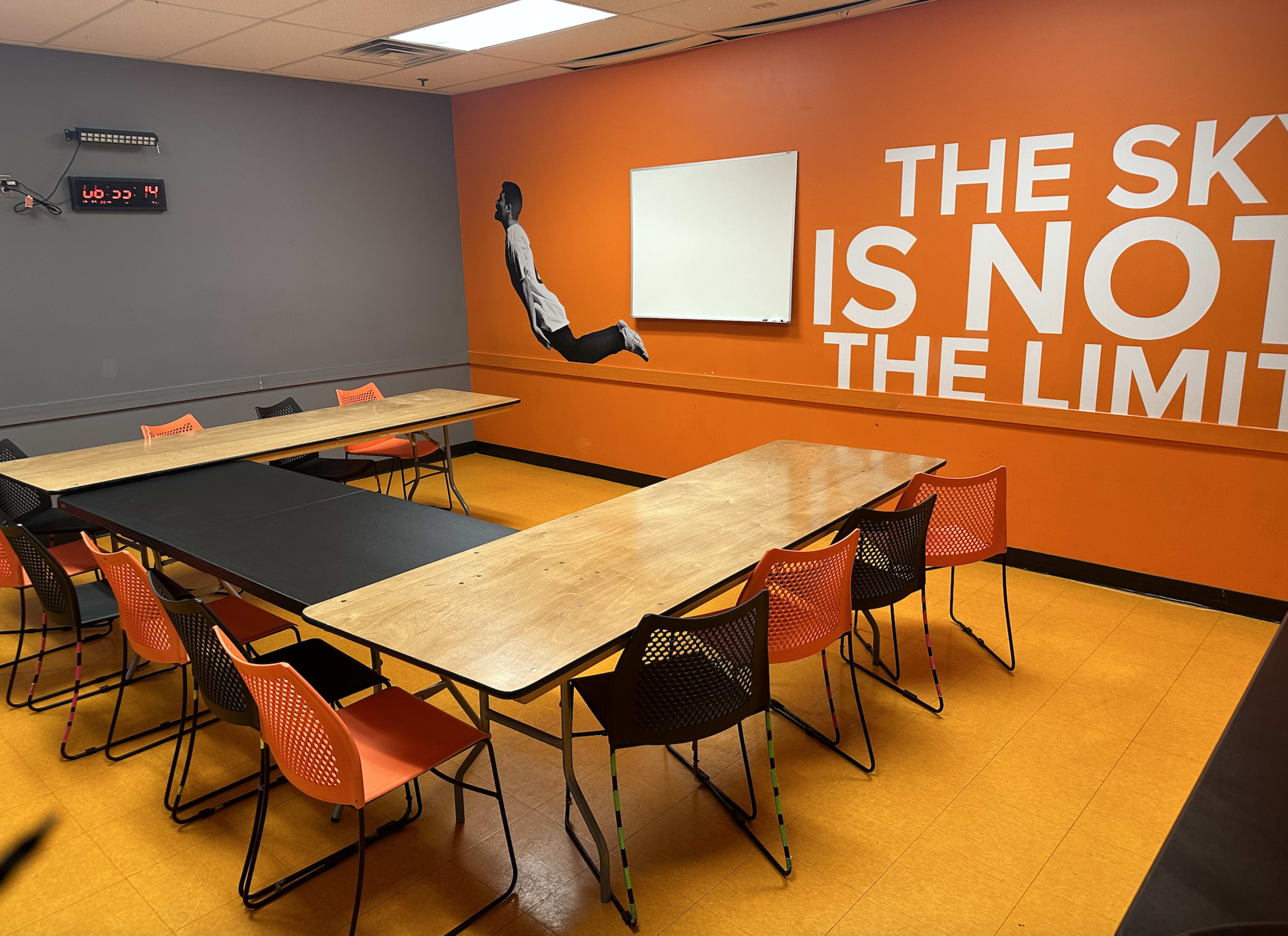 A meeting room features two tables arranged in an L-shape, surrounded by orange and black chairs, with an inspirational wall mural and a whiteboard.