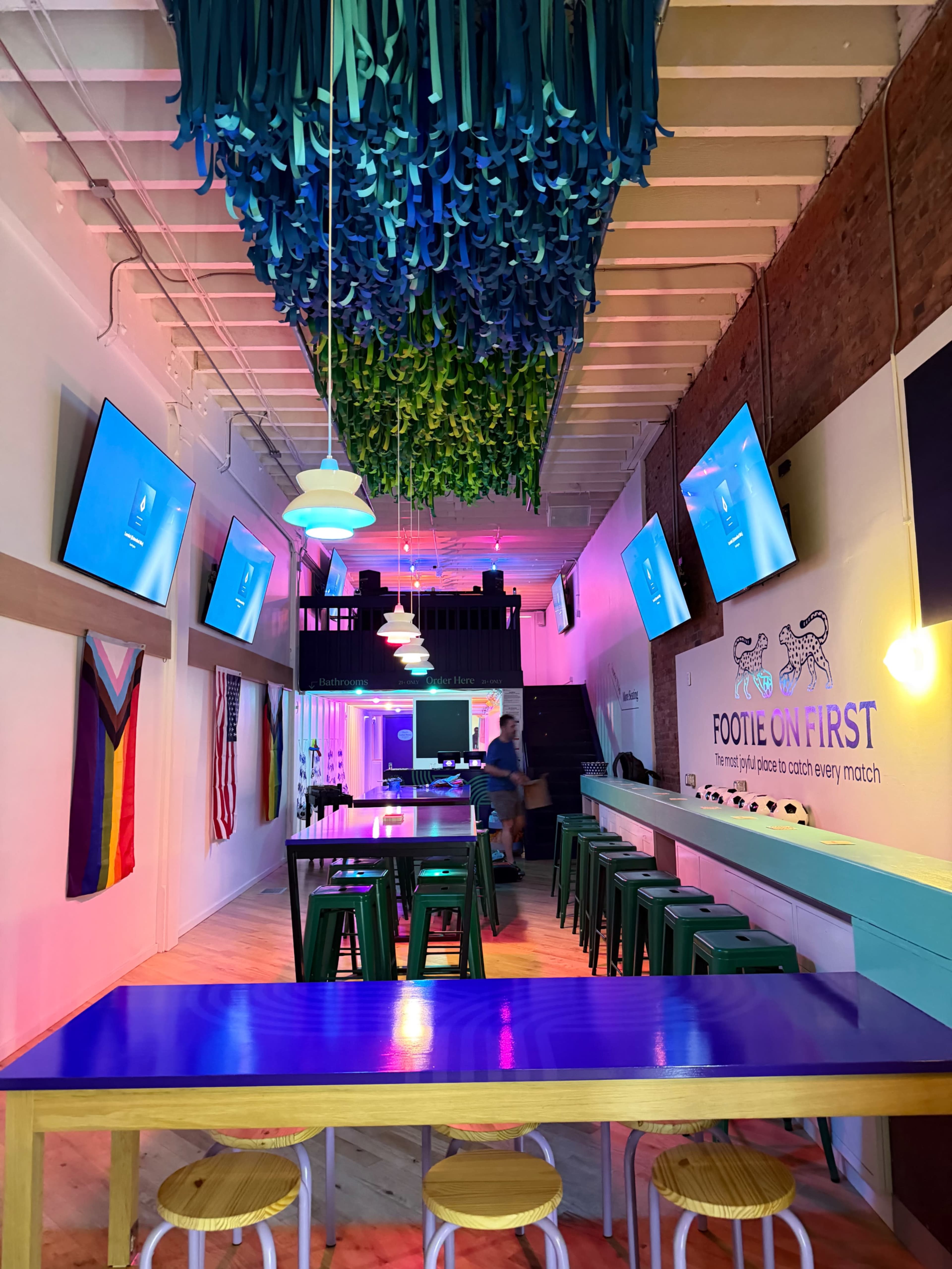 The image shows a vibrant bar interior with colorful overhead decorations, multiple screens displaying content, and a long table lined with green stools.