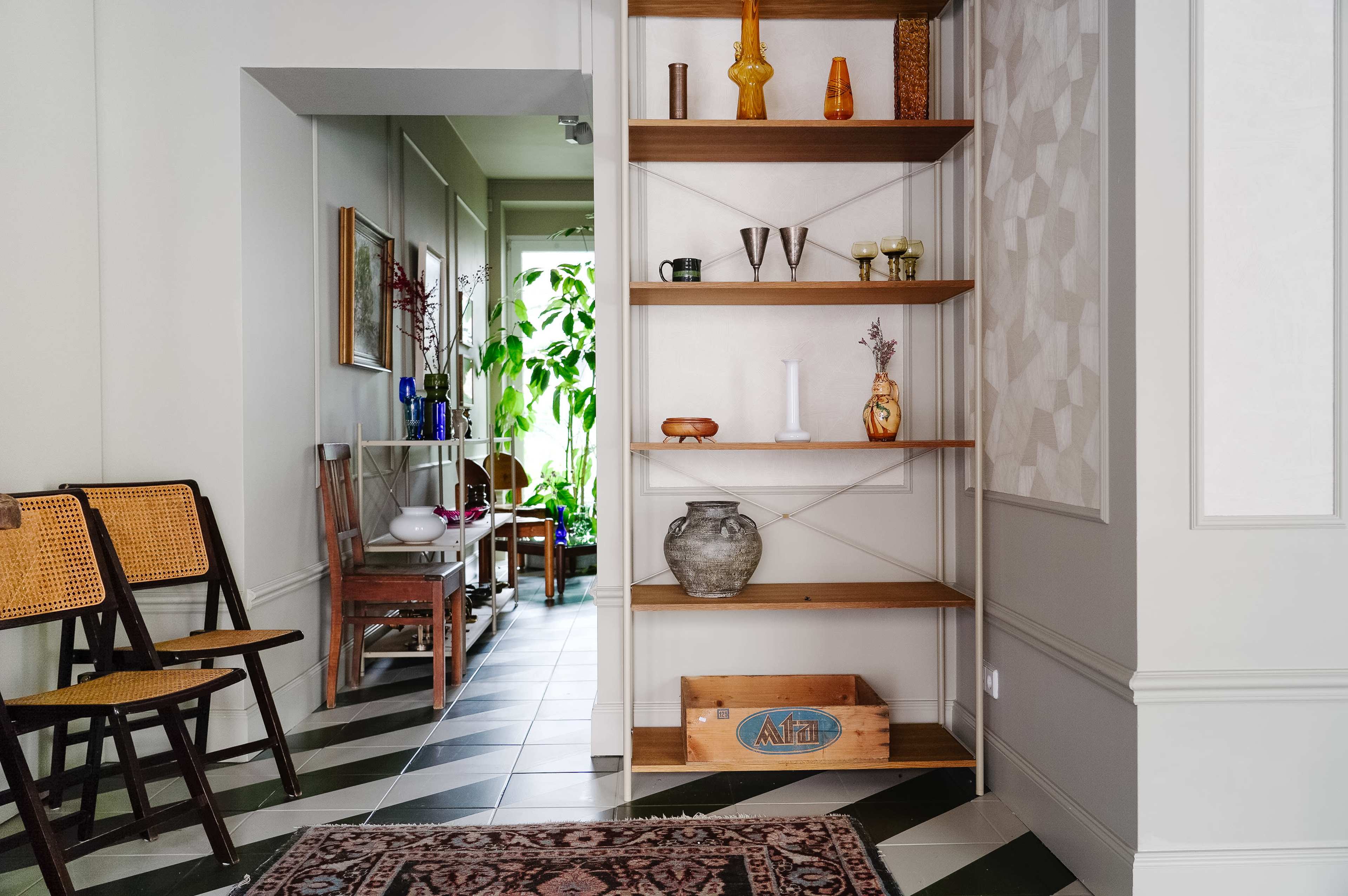 The image shows a hallway with a wooden shelf unit displaying decorative items, flanked by chairs and leading to a room filled with plants and furniture.