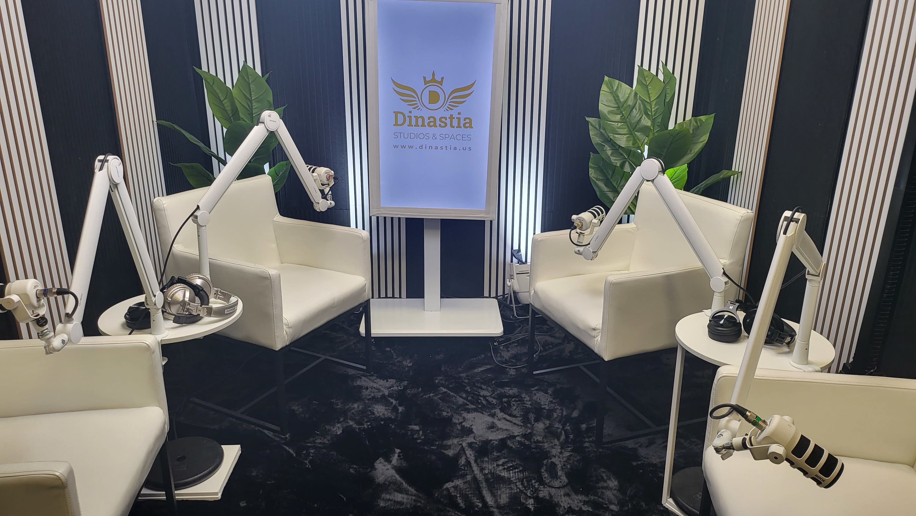The image shows a modern podcast recording studio with white chairs, microphone setups, and decorative plants against a striped wall.