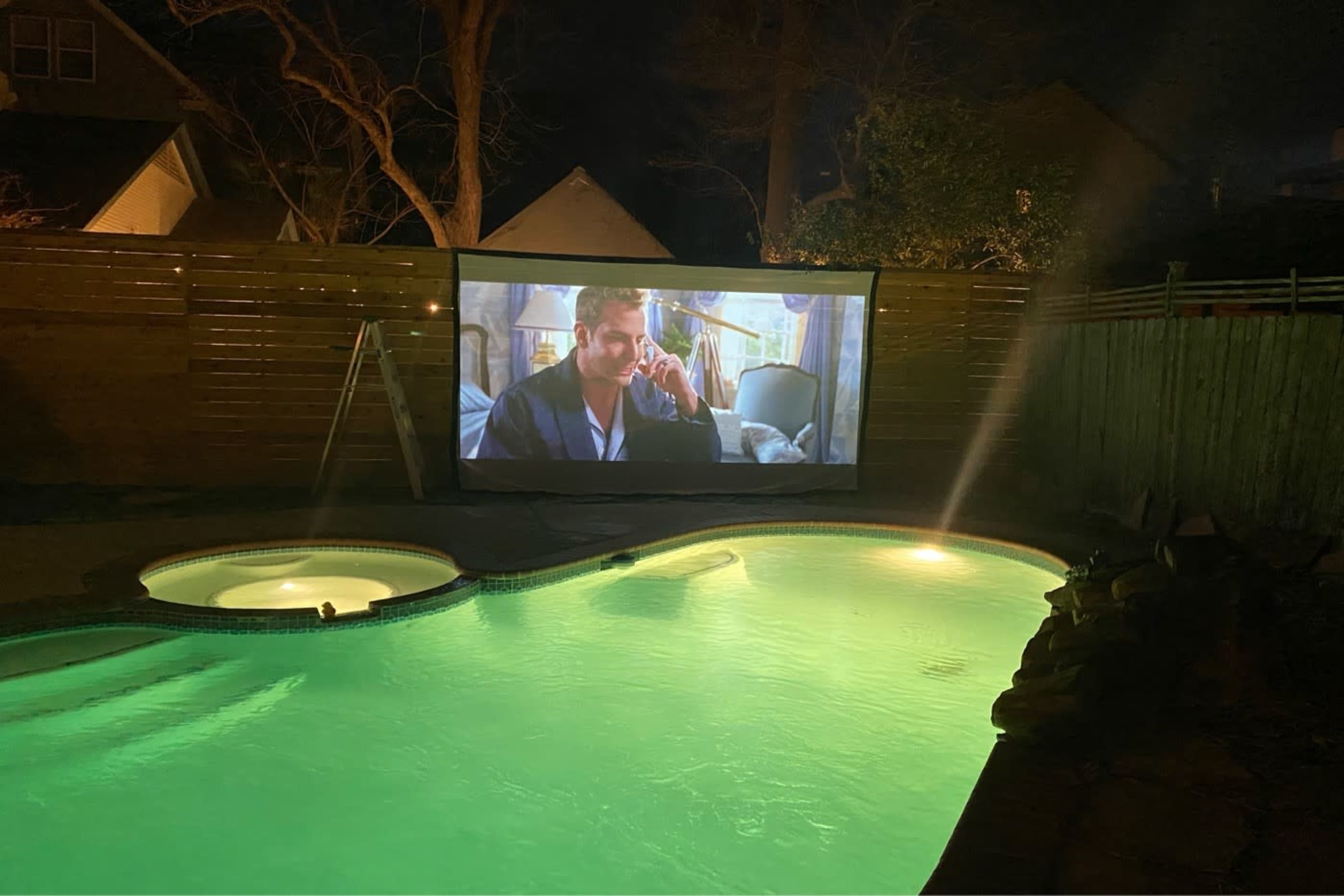 An outdoor movie setup features a large screen displaying a film, illuminated by ambient lighting near a pool with green-tinted water.