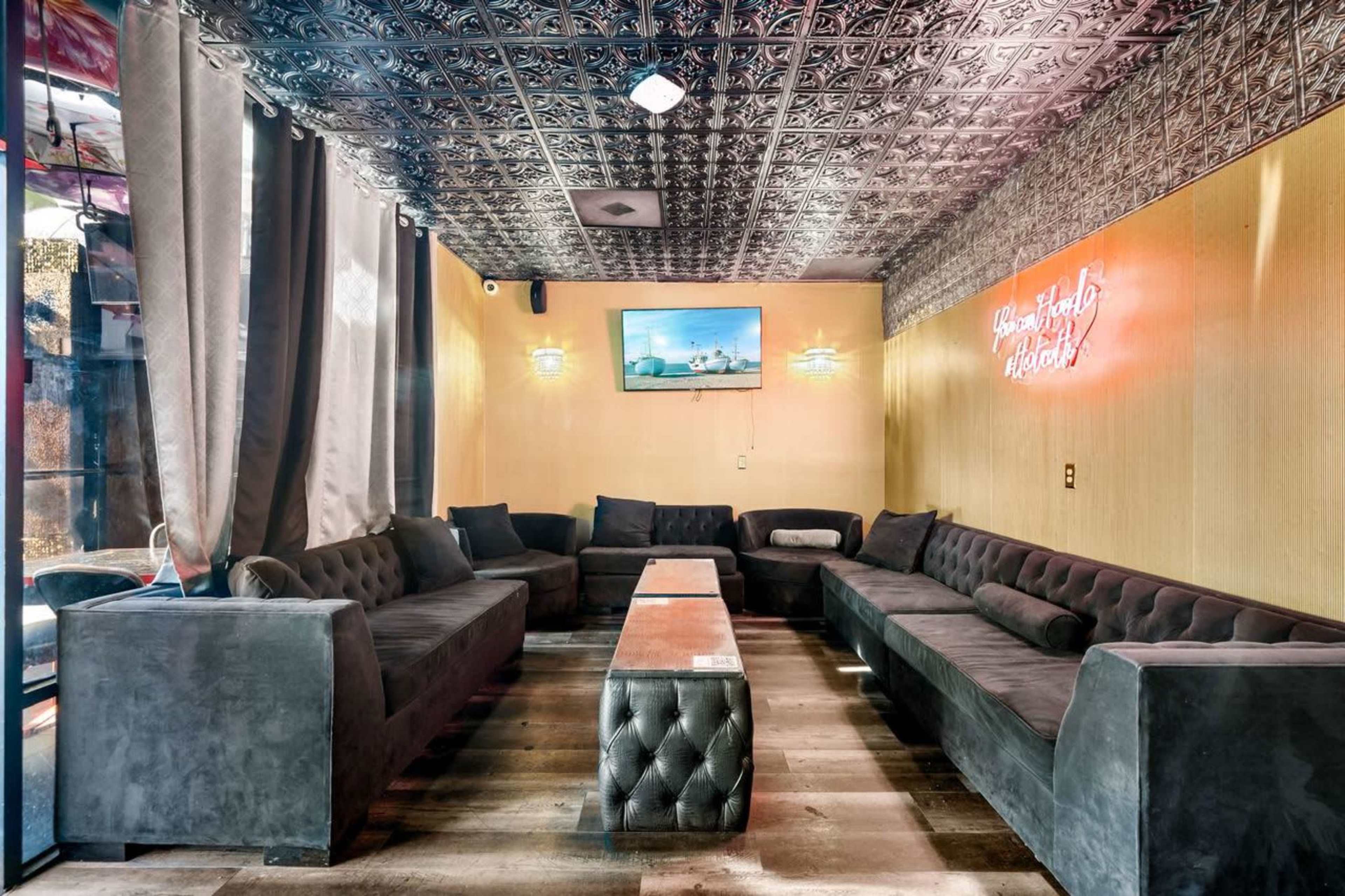 A spacious lounge area features dark velvet seating arranged around a central table, with a decorative ceiling and a wall-mounted television.