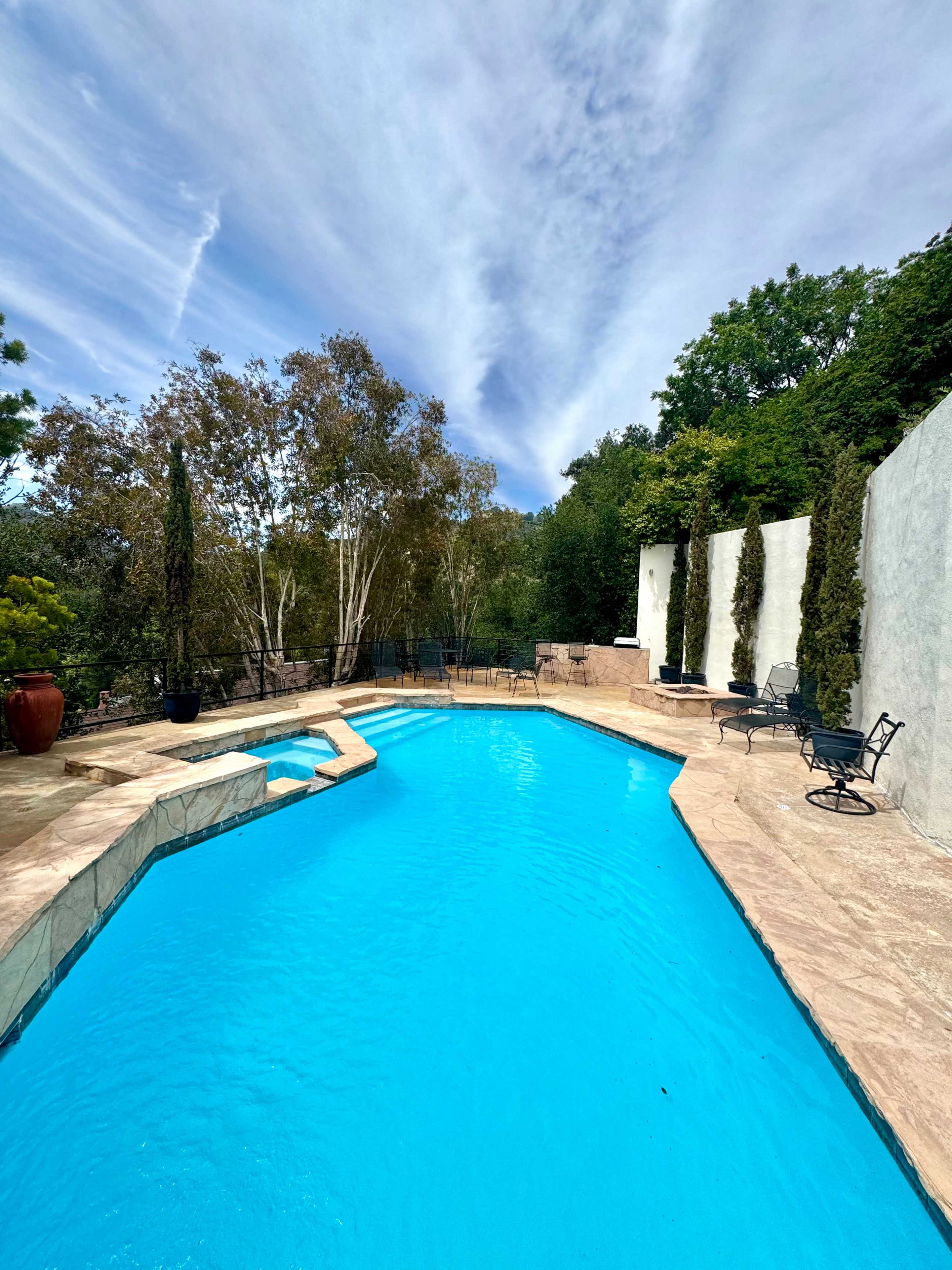 The image shows a clear blue swimming pool surrounded by a stone patio, greenery, and comfortable seating areas.