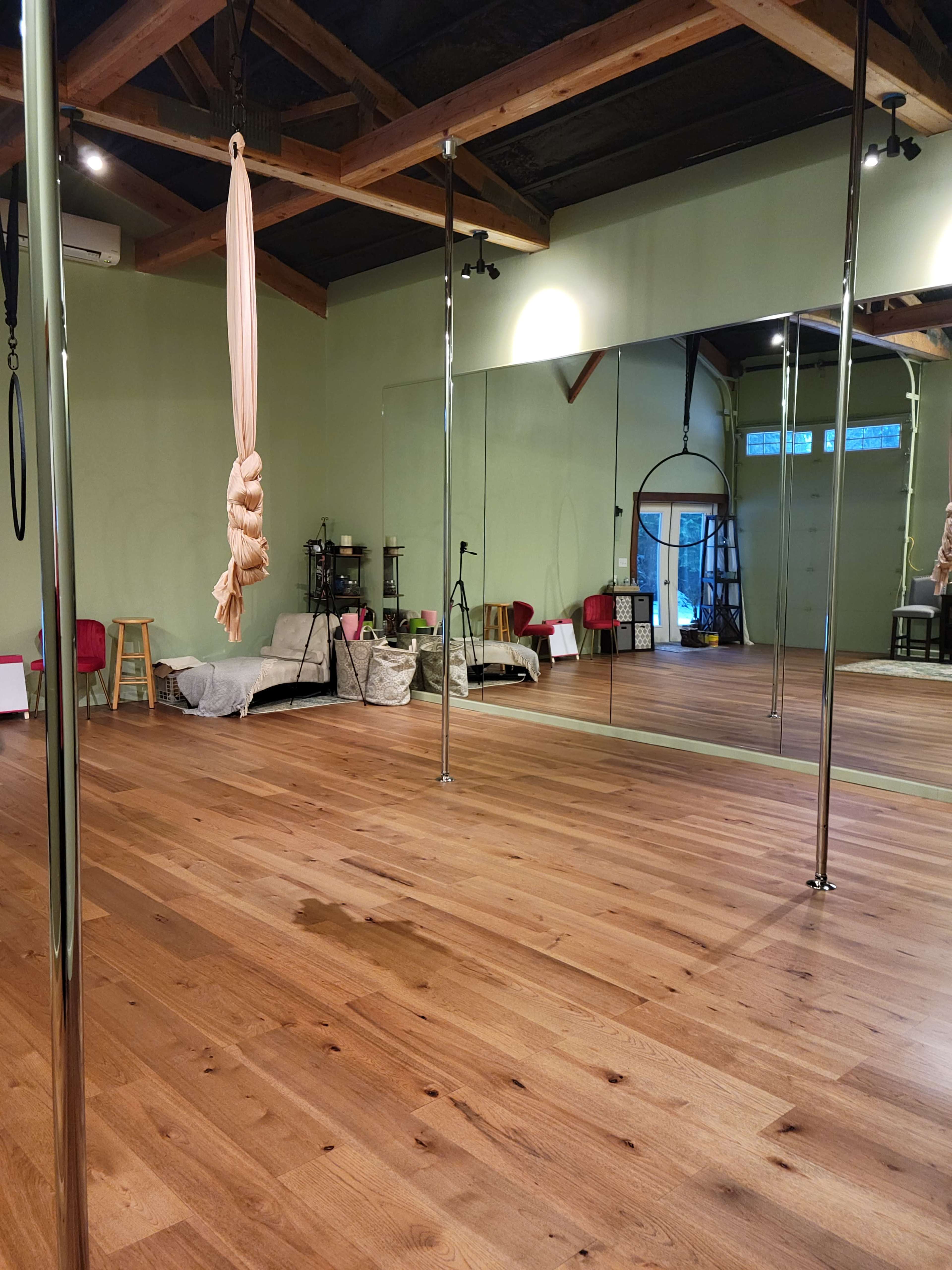 The image shows an empty dance studio with wooden flooring, mirrors on one wall, a silk rope hanging from the ceiling, and various exercise equipment arranged around the space.
