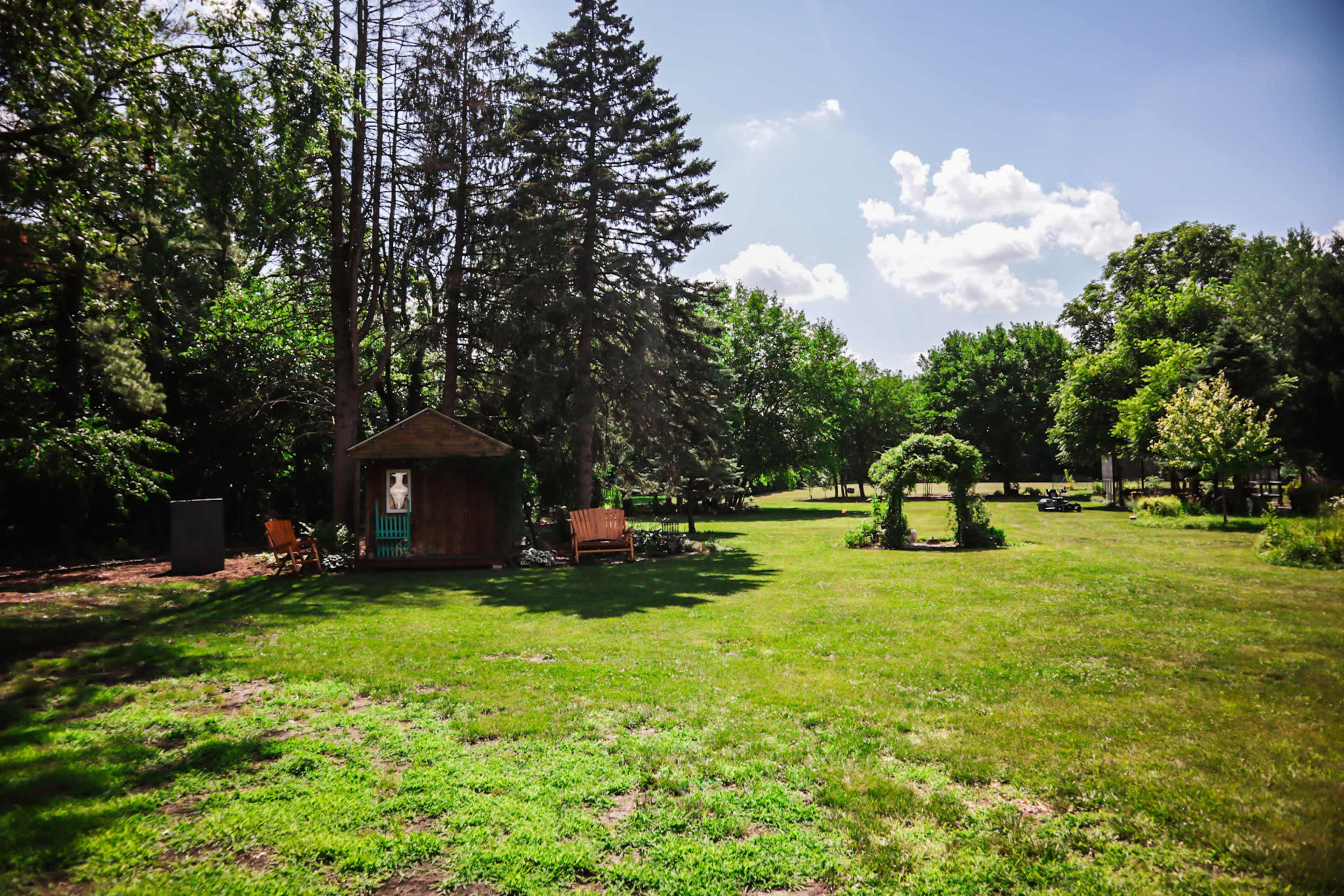 The image shows a grassy park area with a wooden shed, benches, trees, and a decorative archway.