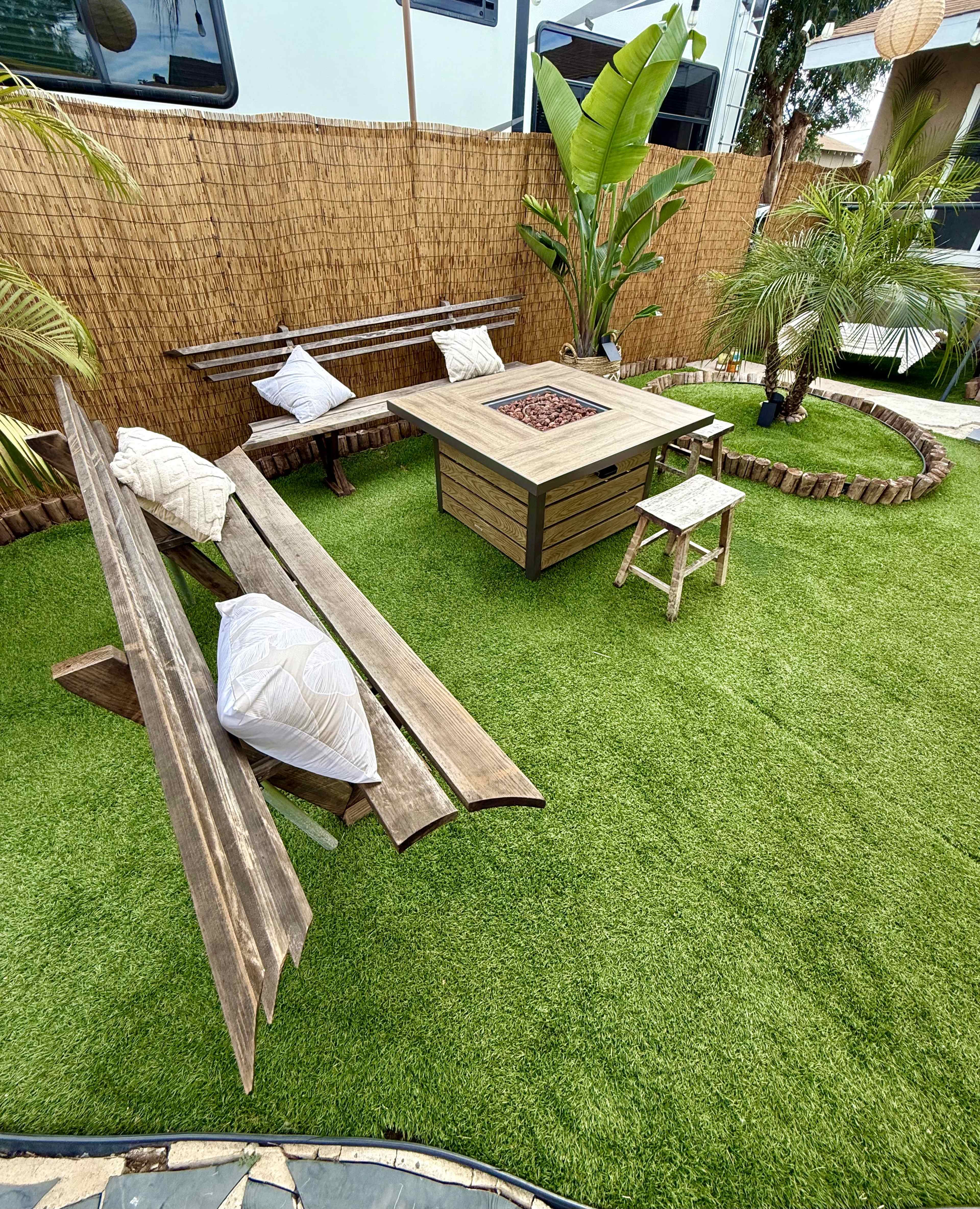 The image shows a landscaped outdoor area featuring a wooden seating arrangement, a fire pit table at the center, and lush green artificial grass surrounded by plants.