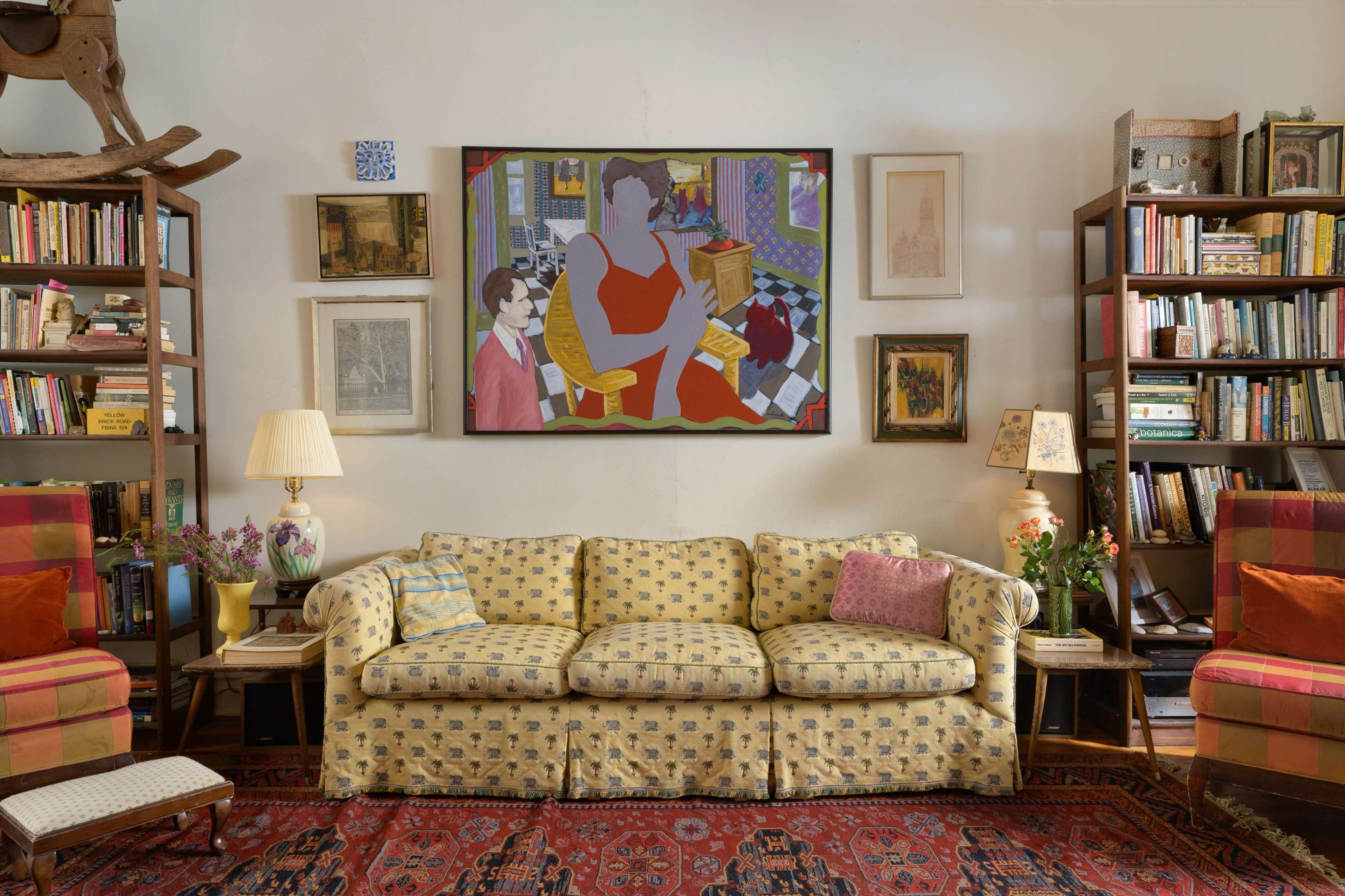 The image shows a brightly decorated living room with a patterned couch, bookshelves filled with books, and a large painting on the wall above the couch.