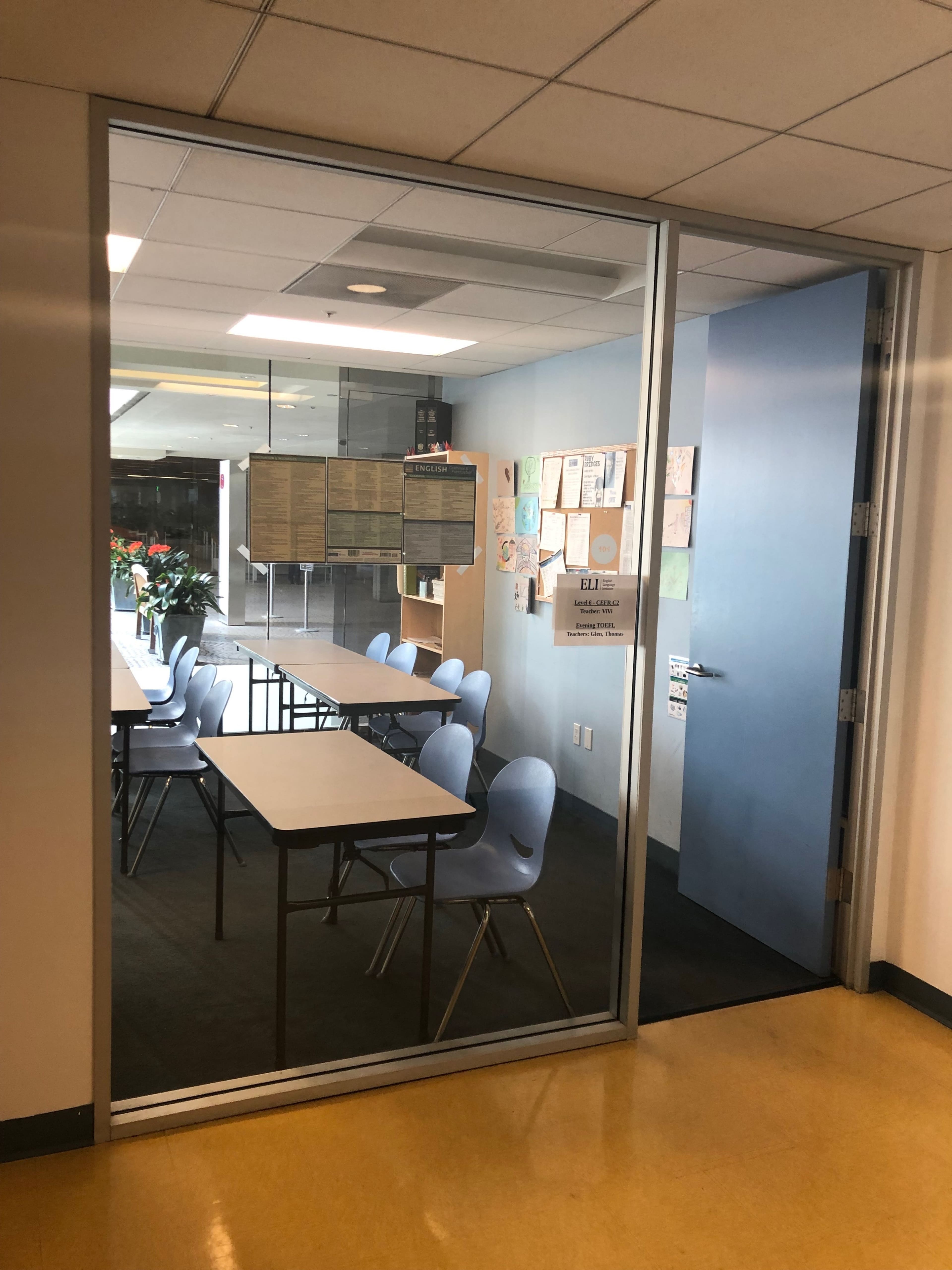 A classroom with several blue chairs and tables, visible through a glass door, with posters and documents on the walls.