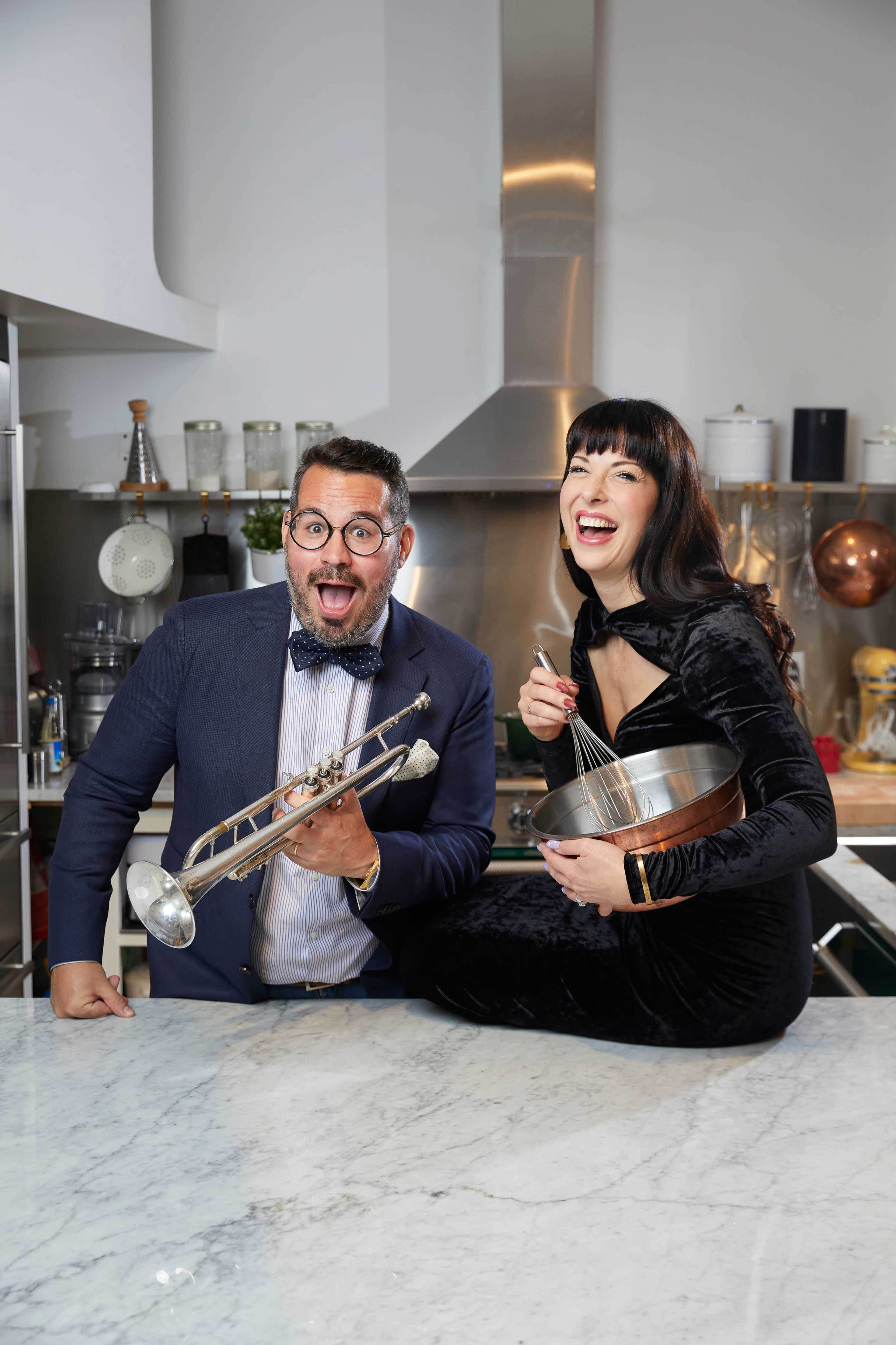 A man holding a trumpet and a woman holding a mixing bowl and whisk are smiling and posing on a marble countertop in a kitchen.