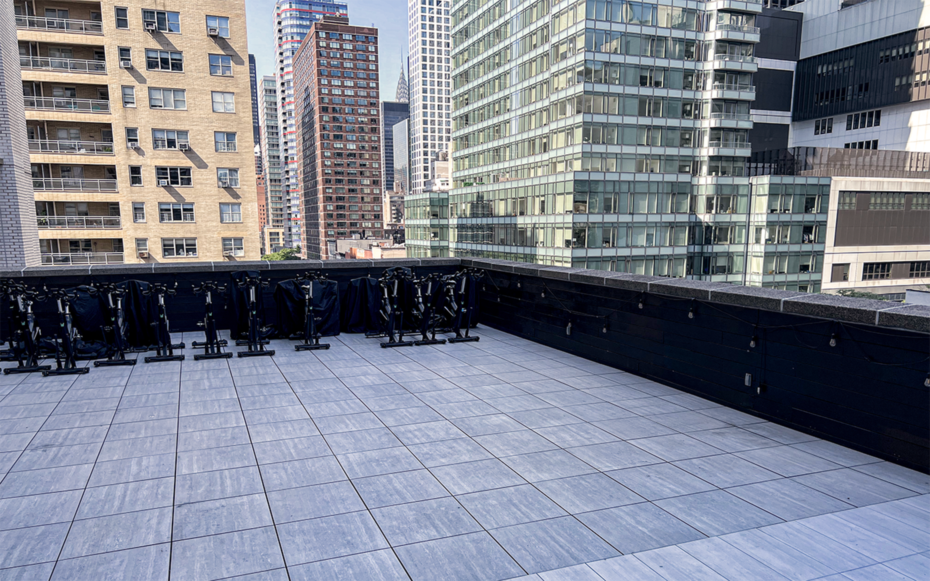 A rooftop terrace features a tiled floor and a row of black clothing racks against a backdrop of tall buildings.