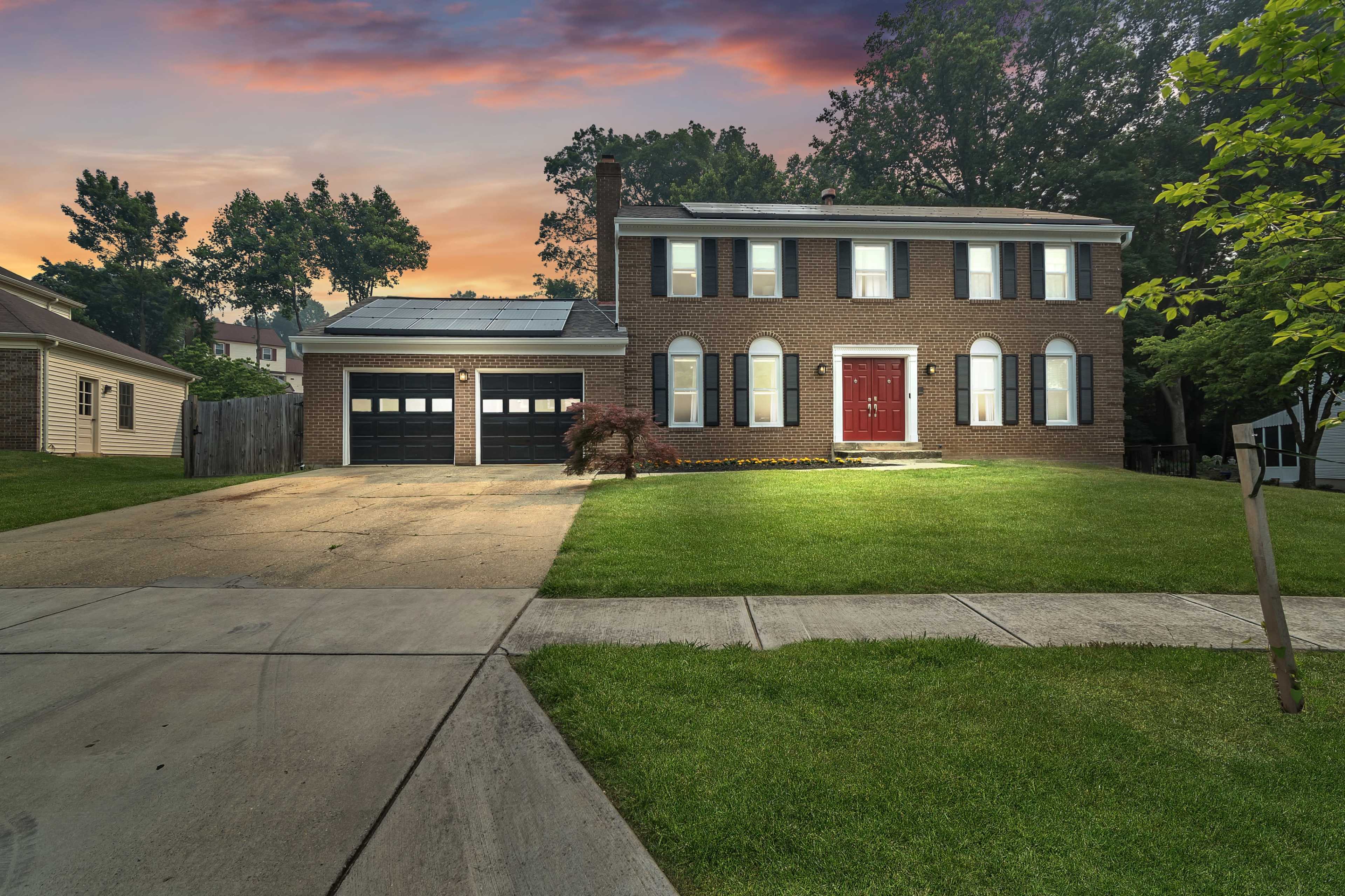 A two-story brick house with a red front door and solar panels on the roof sits on a grassy lot with a driveway and two garage doors.