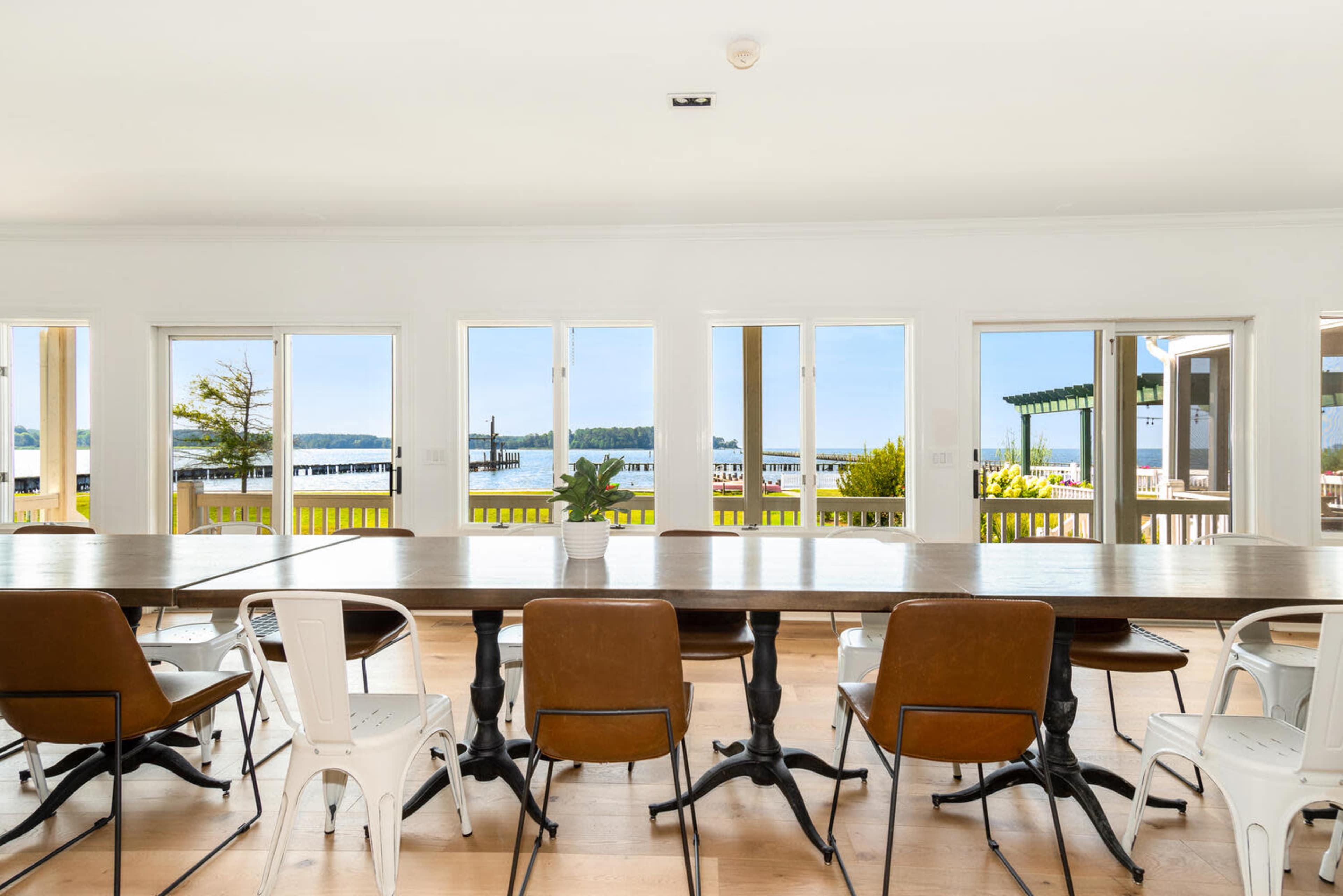 A long wooden table with metal and leather chairs is positioned beside large windows overlooking a lake and docks.