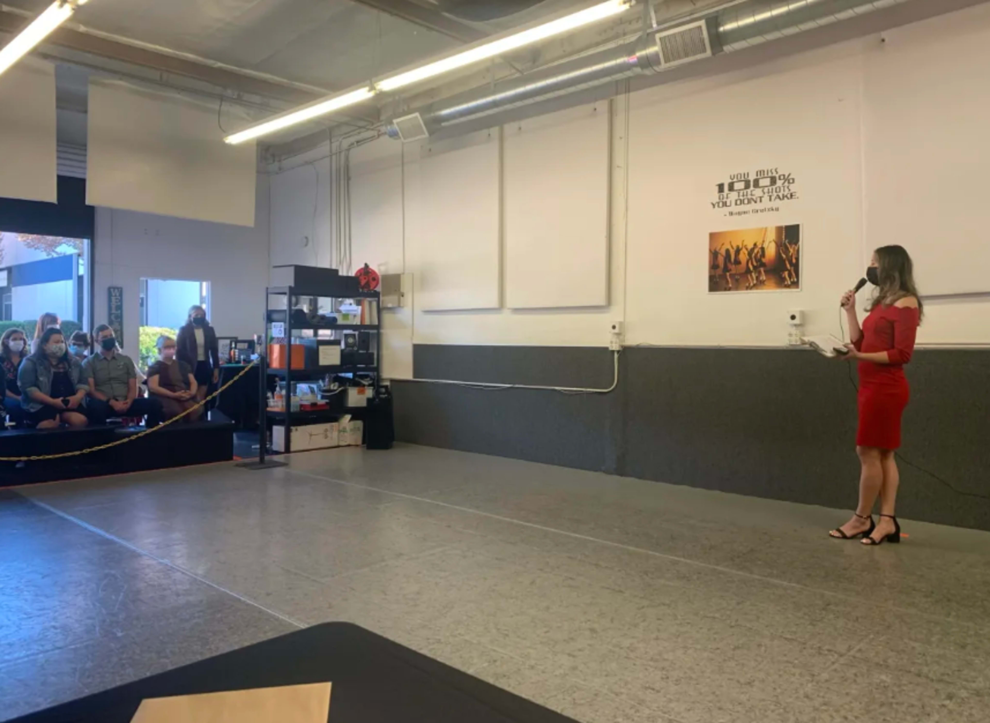 A woman in a red dress speaks to an audience seated in a studio space with a large mirror and shelves of supplies in the background.