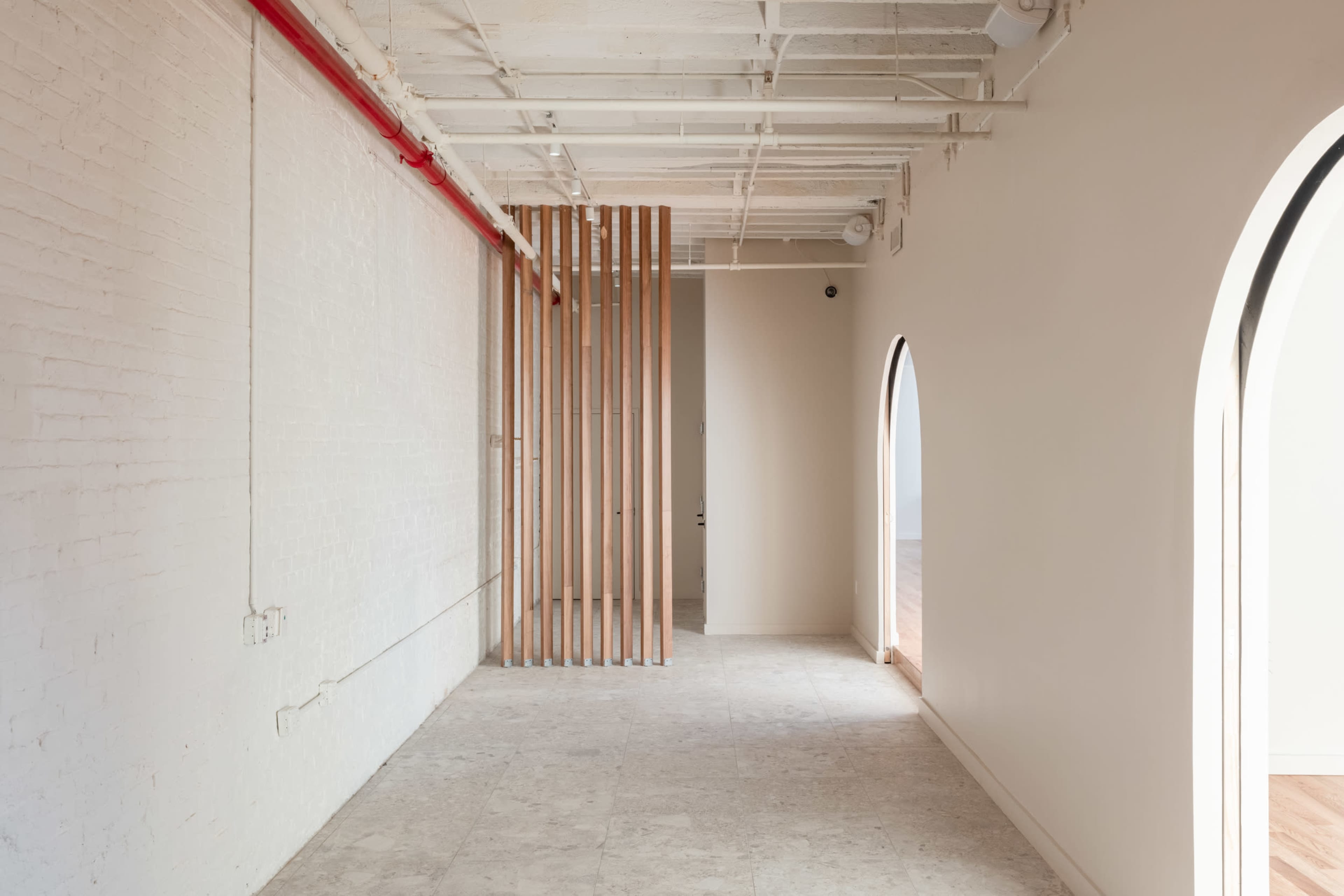 The image shows a modern, minimalist interior space with wooden slats dividing the area and arched openings on one side.