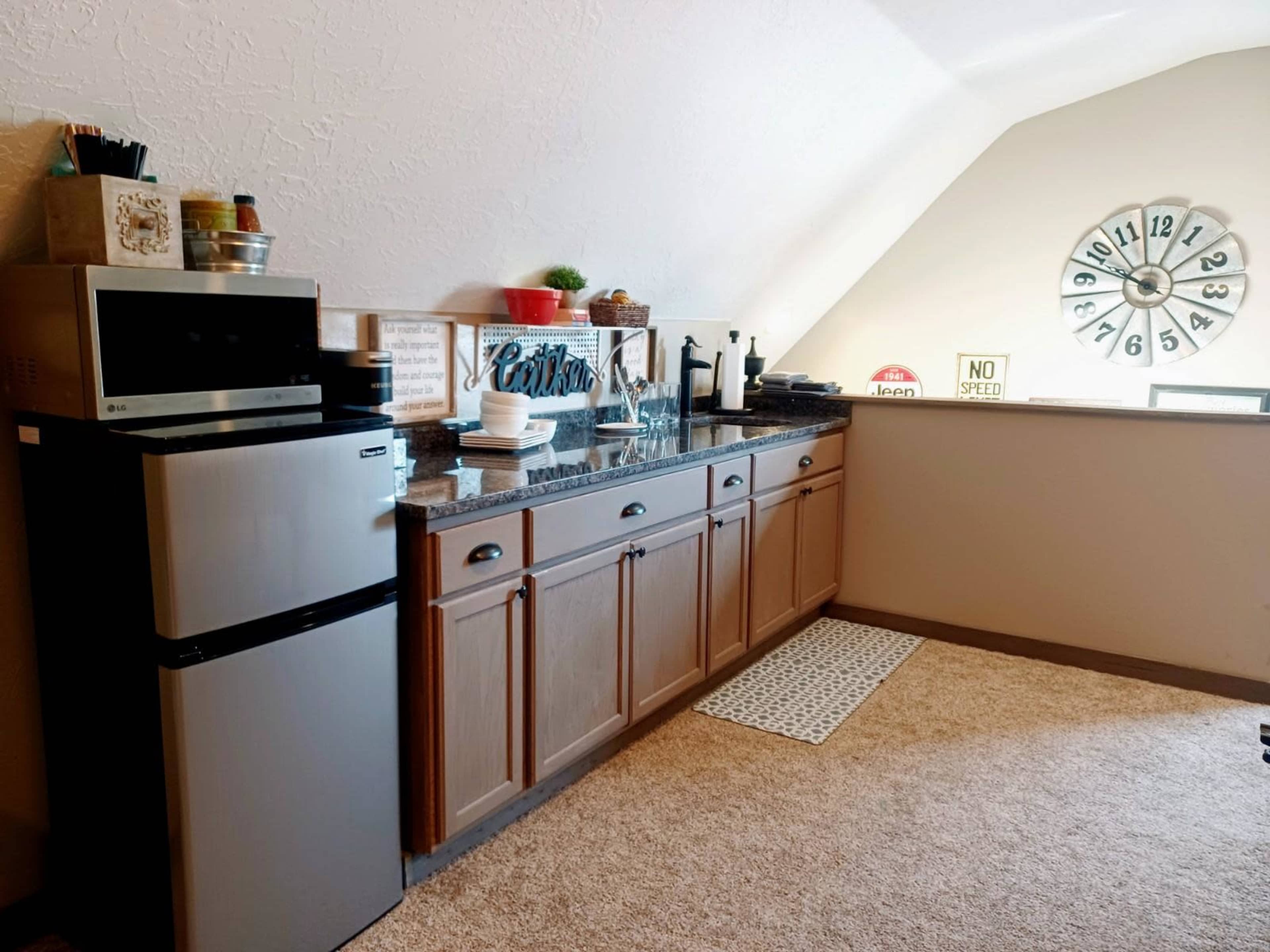 A kitchenette features a small refrigerator, granite countertop, and a clock on the wall with an angled ceiling above.