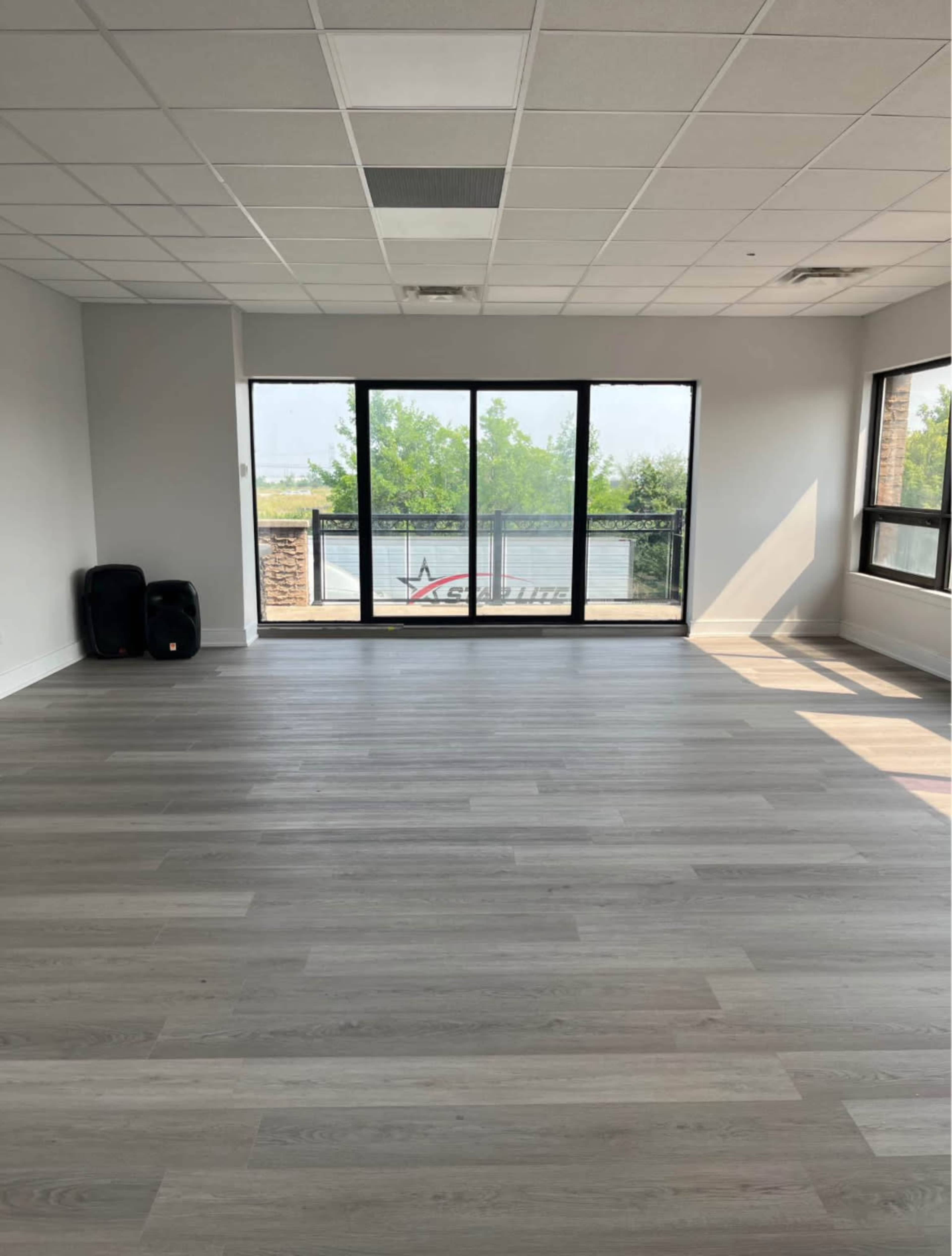 The image shows a spacious room with light-colored wooden flooring and large windows overlooking a green landscape.