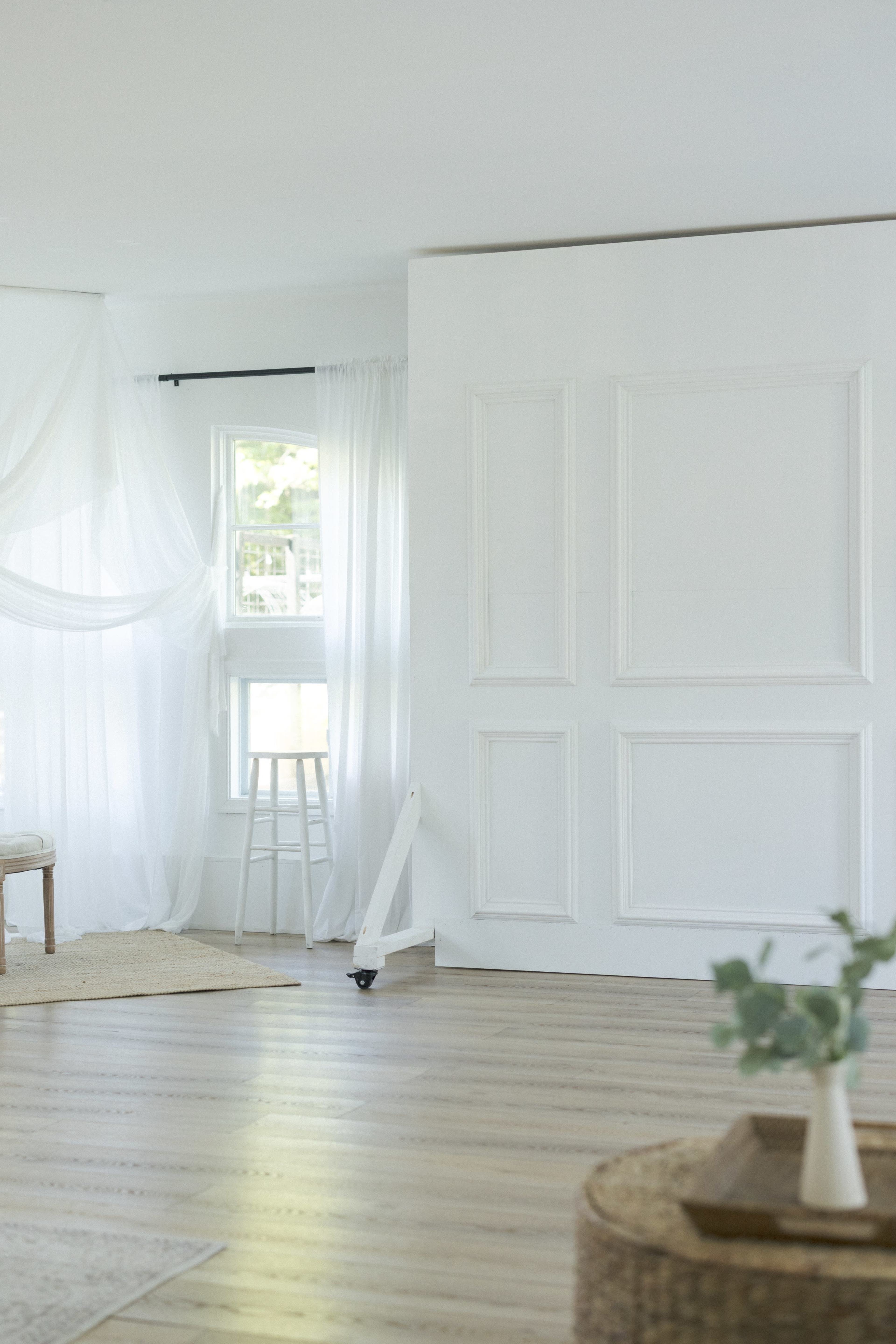 The image features a bright, minimalist room with white walls, sheer curtains, and wooden flooring.