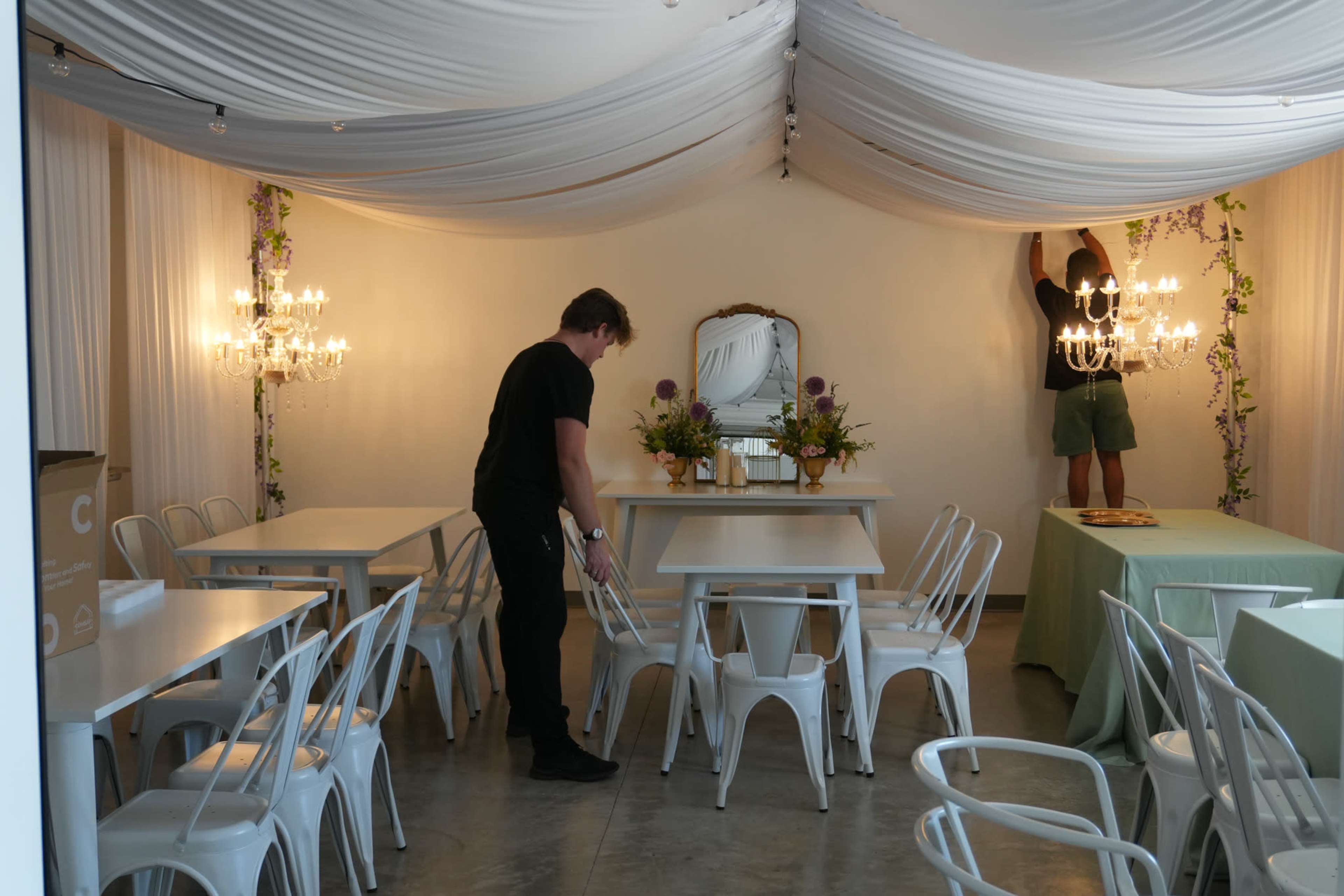 Two individuals are arranging furniture in a well-decorated indoor space with draped fabric and hanging chandeliers.