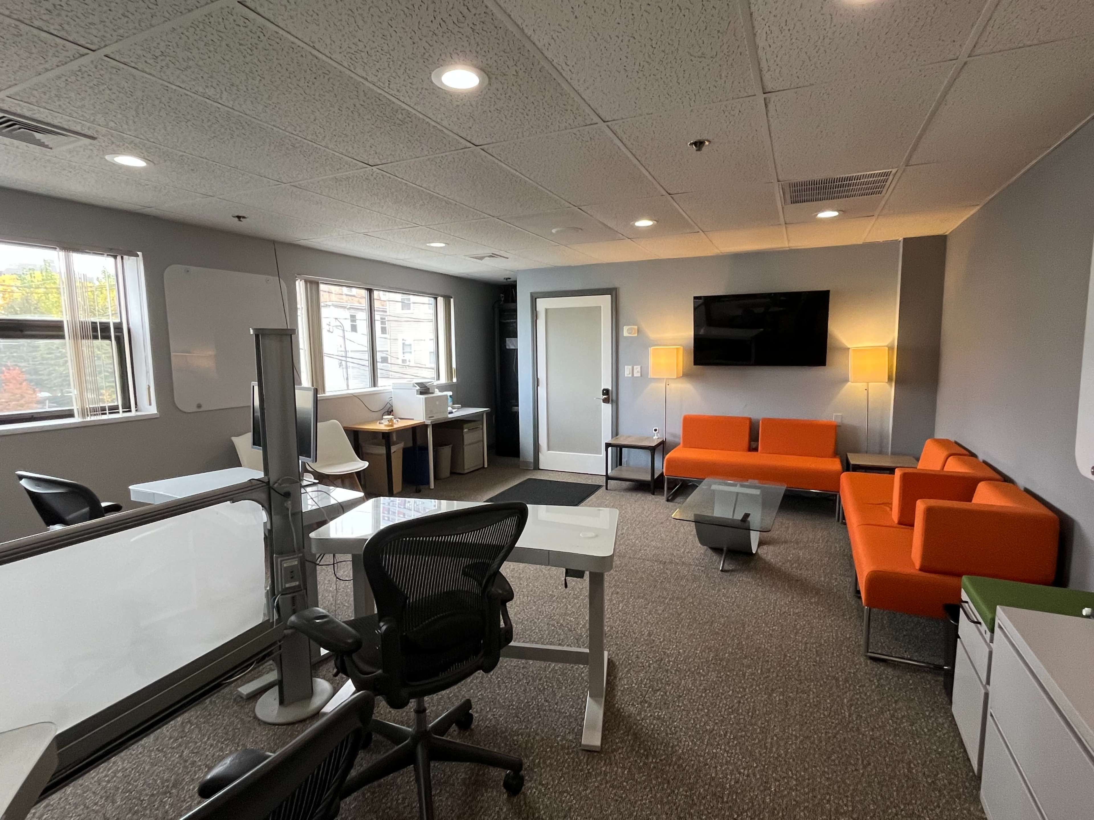 The image shows a modern office space featuring an orange sofa, white desks, and a large television mounted on the wall.