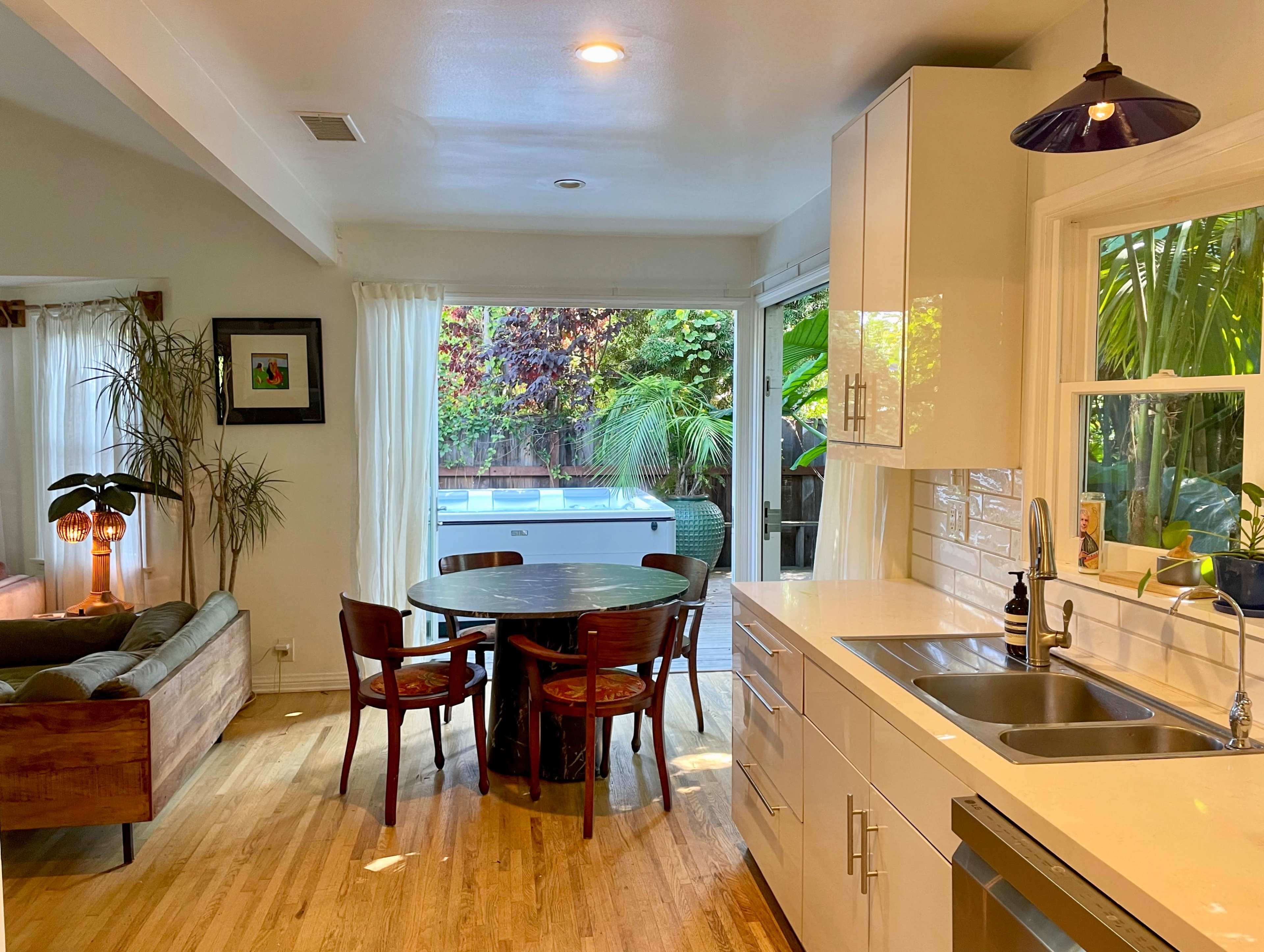 A modern kitchen features a round dining table surrounded by wooden chairs, with large windows providing views of a lush outdoor garden.