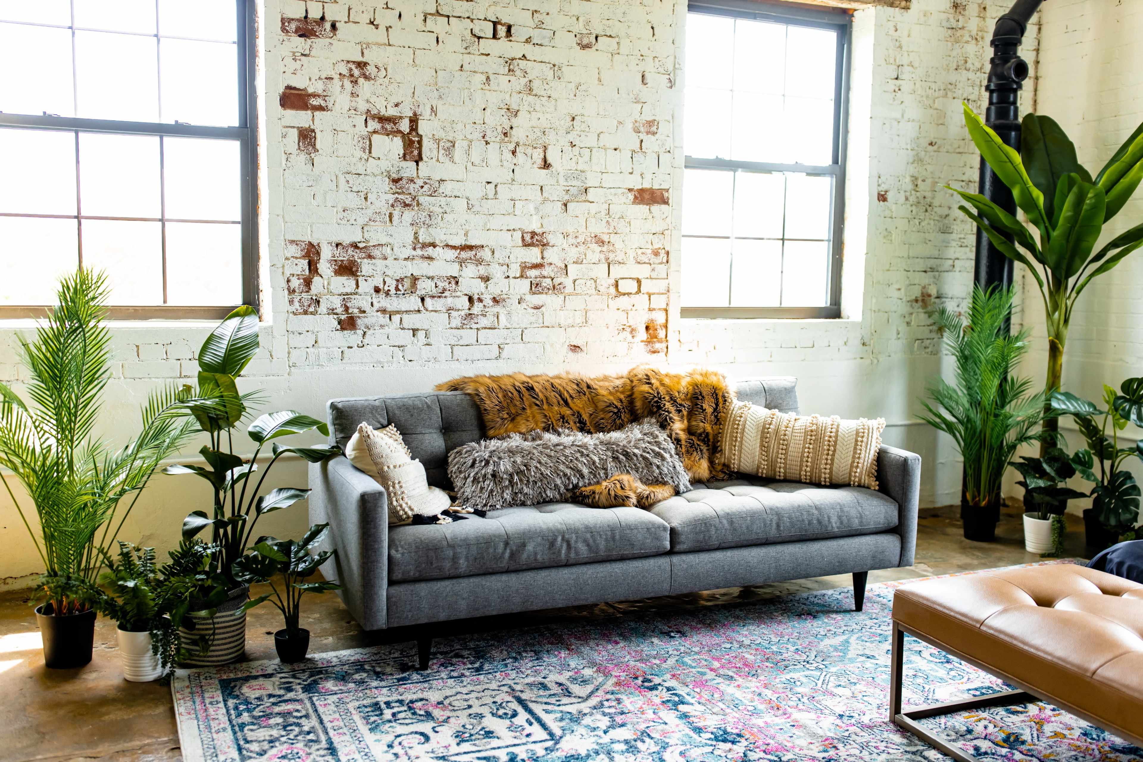 A gray sofa is positioned against a brick wall, surrounded by indoor plants and accented with various throw pillows.