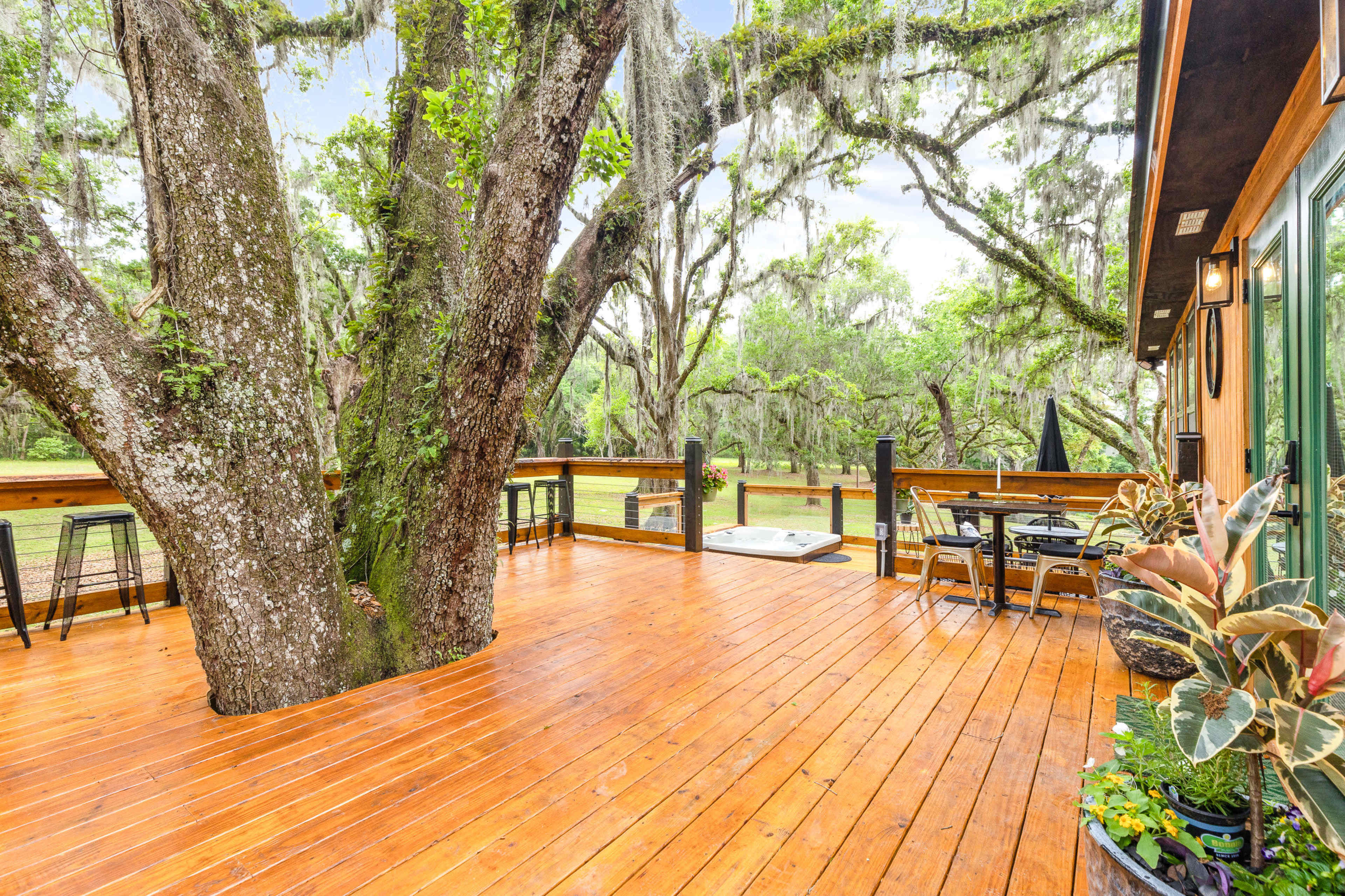 A wooden deck features a large tree at its center, surrounded by outdoor furniture and lush greenery.