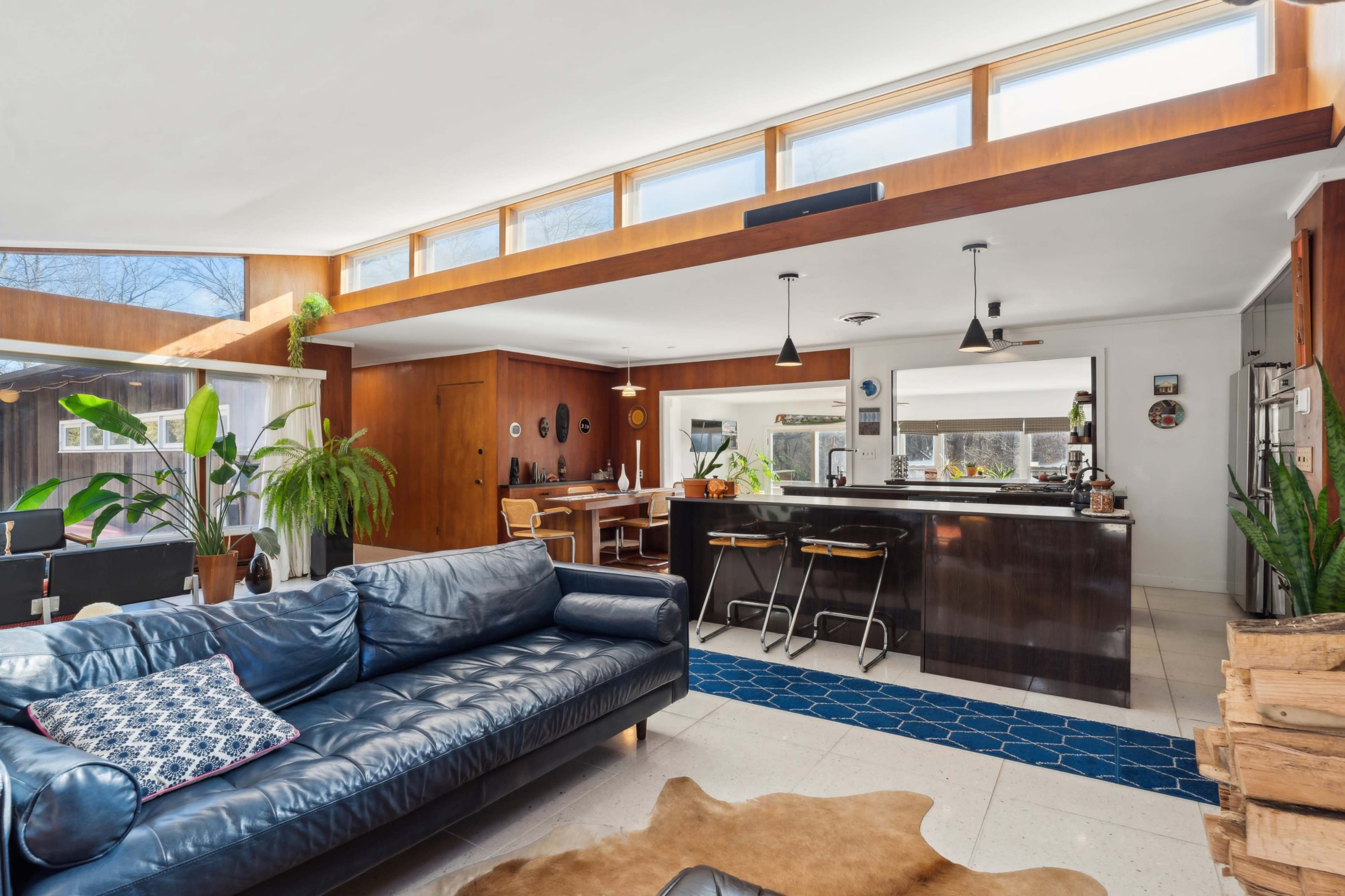 The image shows a modern living space featuring a blue leather sofa, a kitchen with a bar counter, and large windows that allow natural light to illuminate the room.