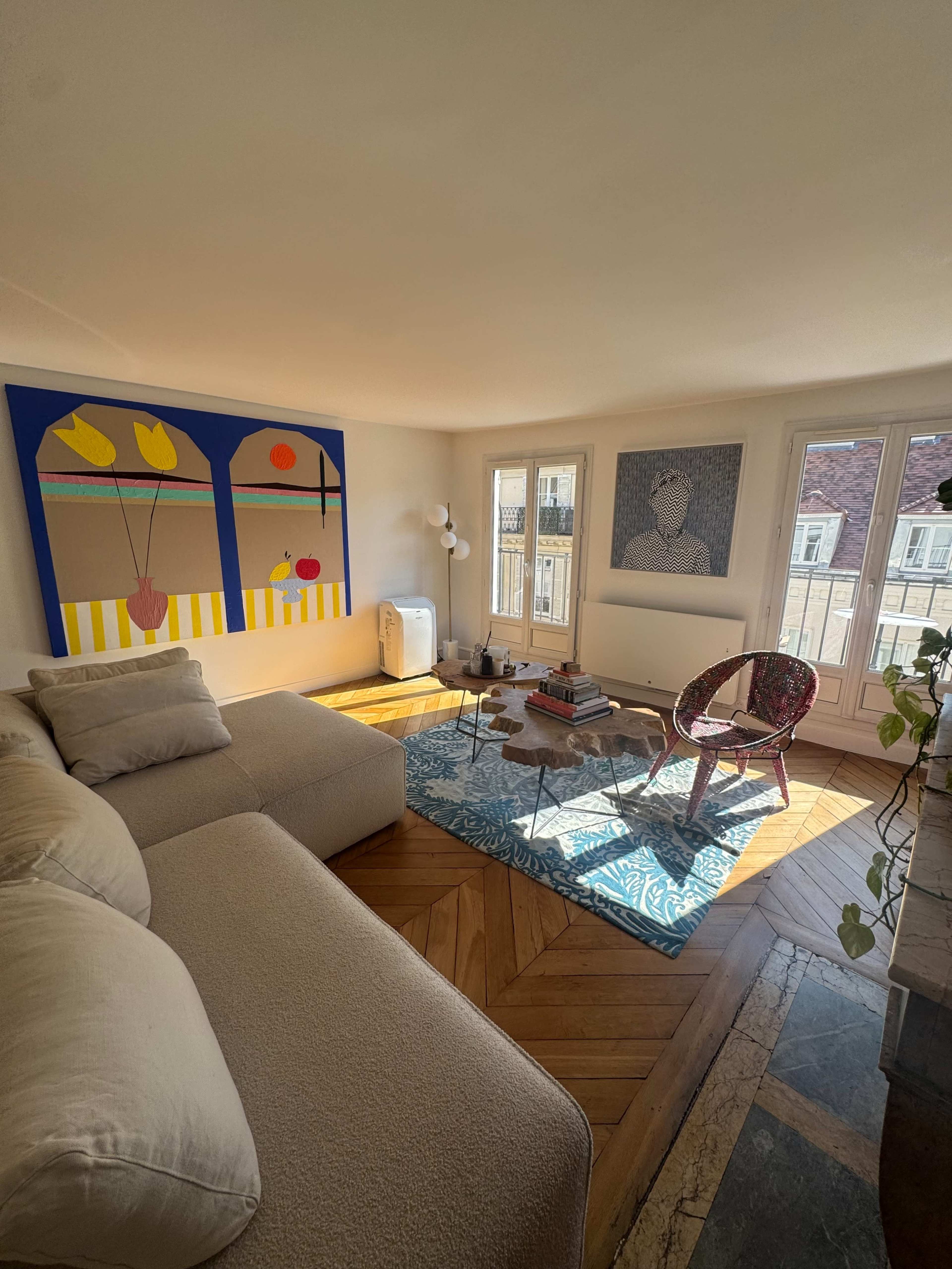 The image shows a bright living room with a large couch, a colorful wall painting, a round coffee table, and large windows letting in natural light.
