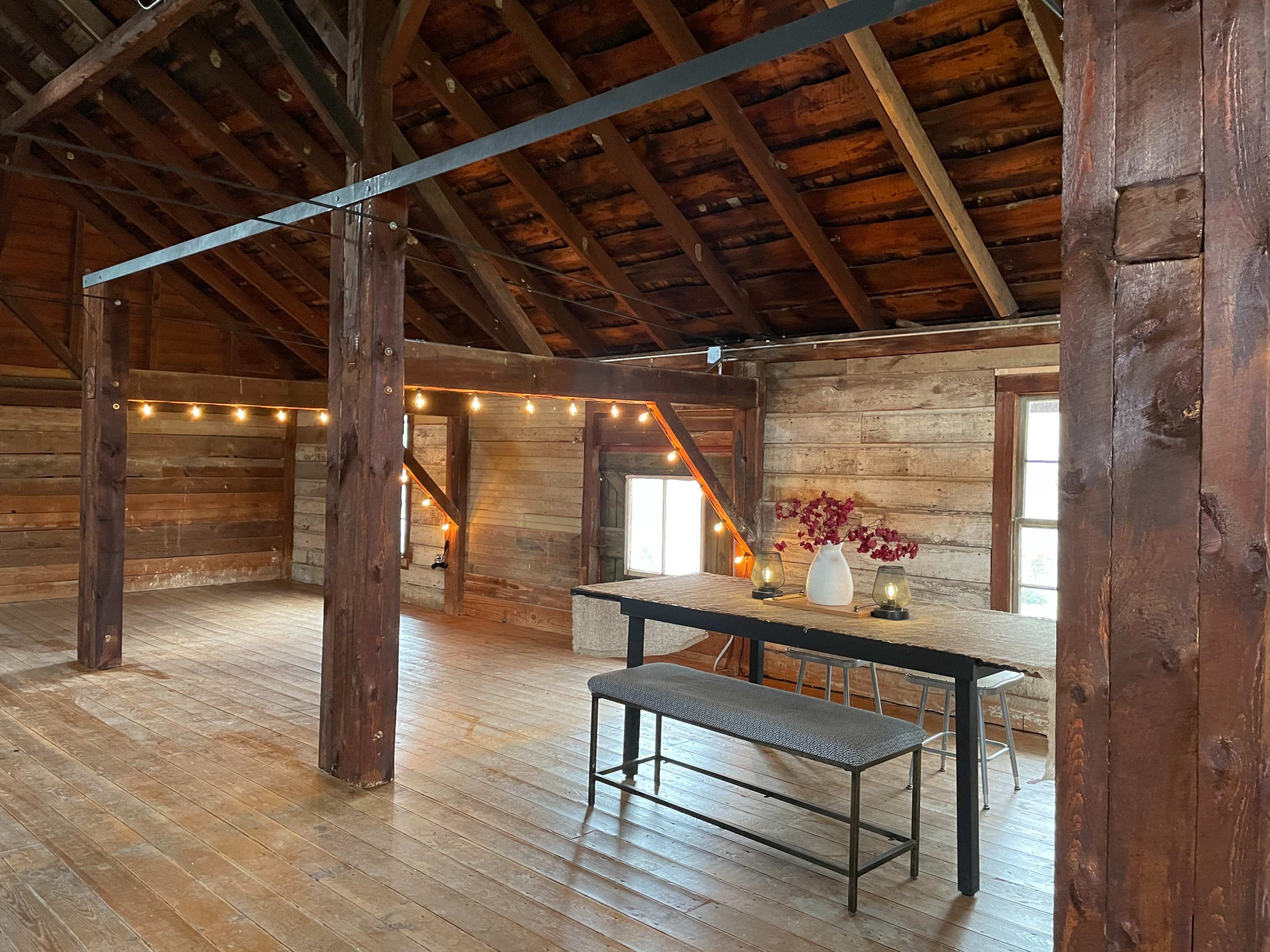 The image shows a rustic, wooden interior of an attic space with exposed beams, a dining table, and a bench, illuminated by string lights.