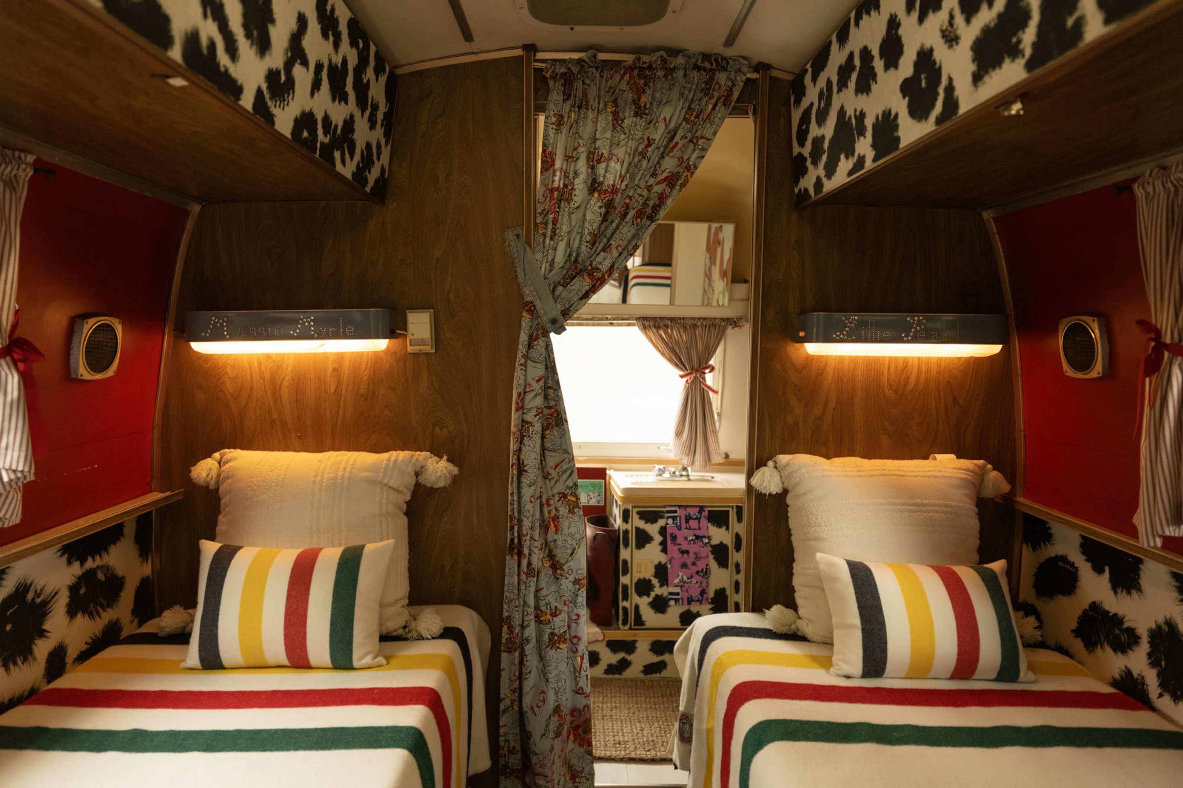 The image shows the interior of a vintage trailer featuring two twin beds with striped blankets, decorative pillows, and cow-print walls.