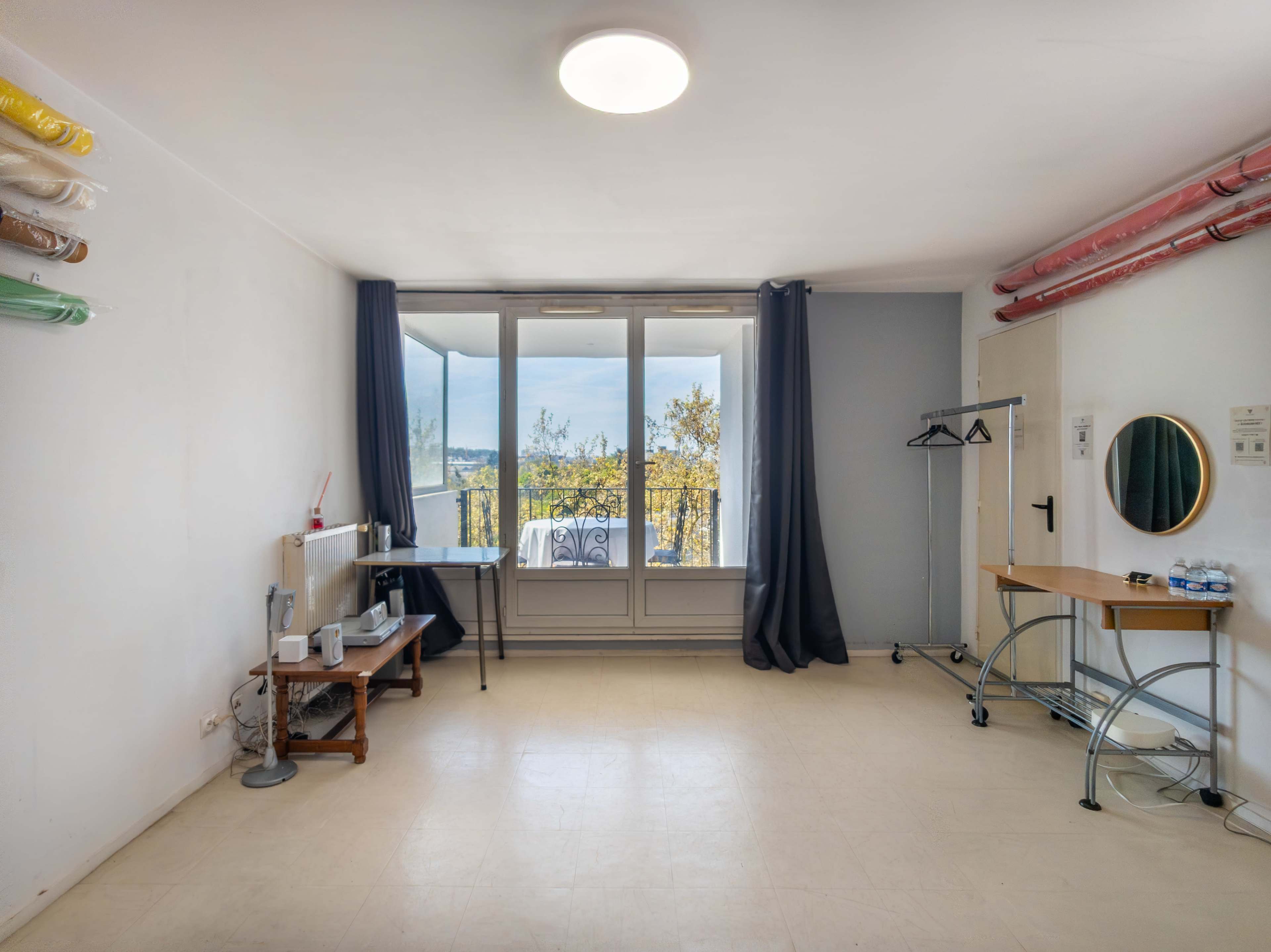 The image shows a spacious, empty room with large windows, a small table and chair, and a coat rack near the wall.