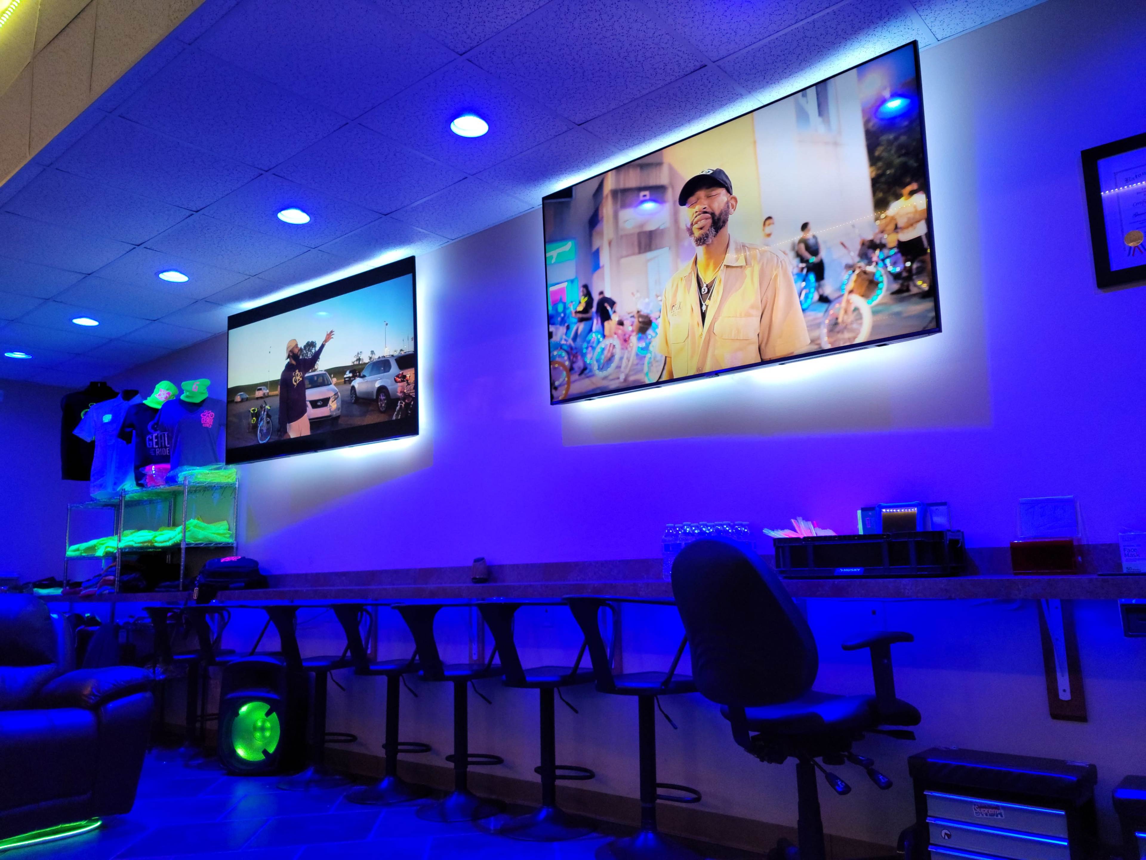 A brightly lit lounge area with two televisions mounted on the wall, displaying videos, and a bar-like seating arrangement with high stools.