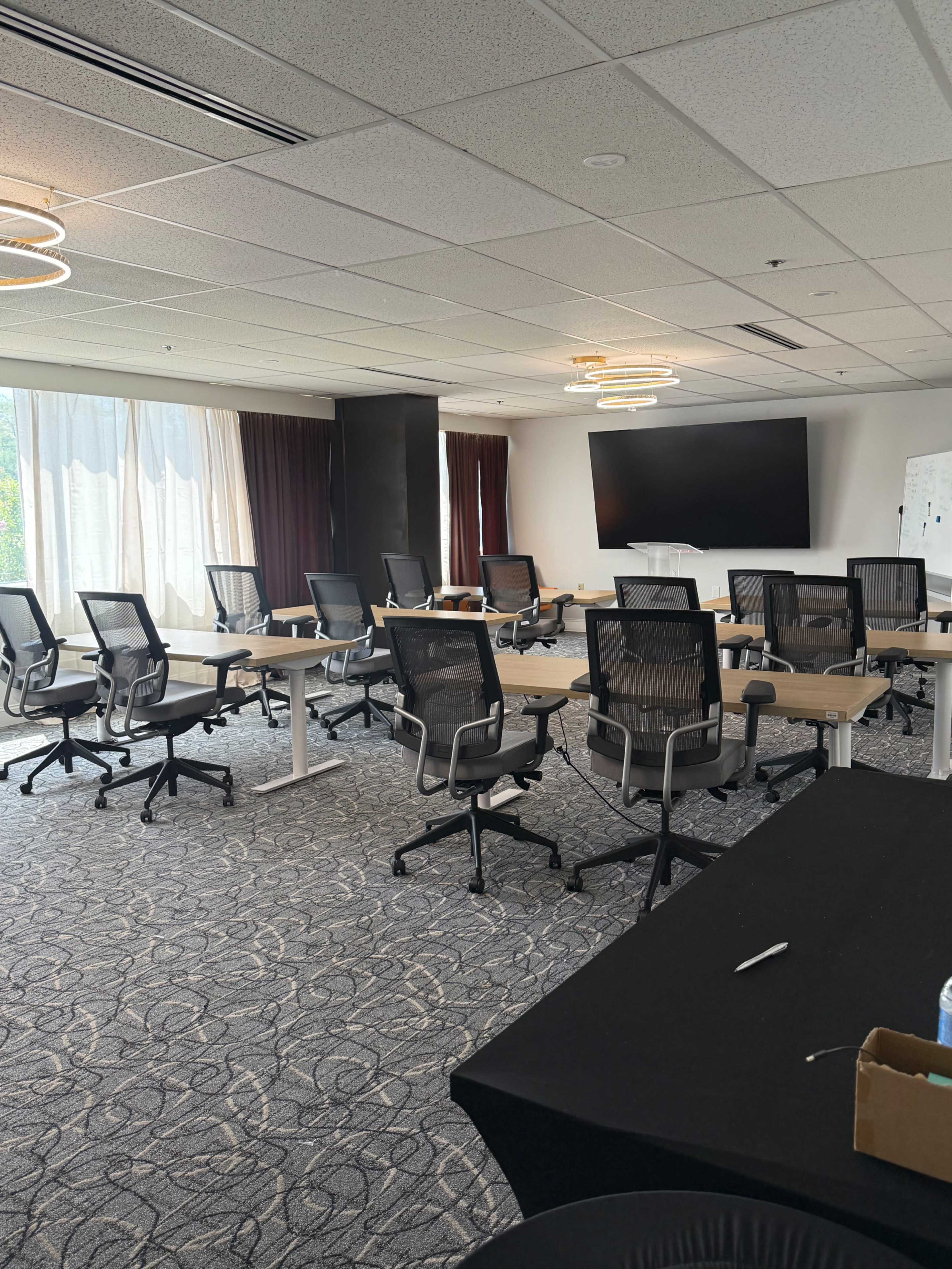 The image shows a modern conference room with several rows of office chairs and tables arranged around a large display screen.