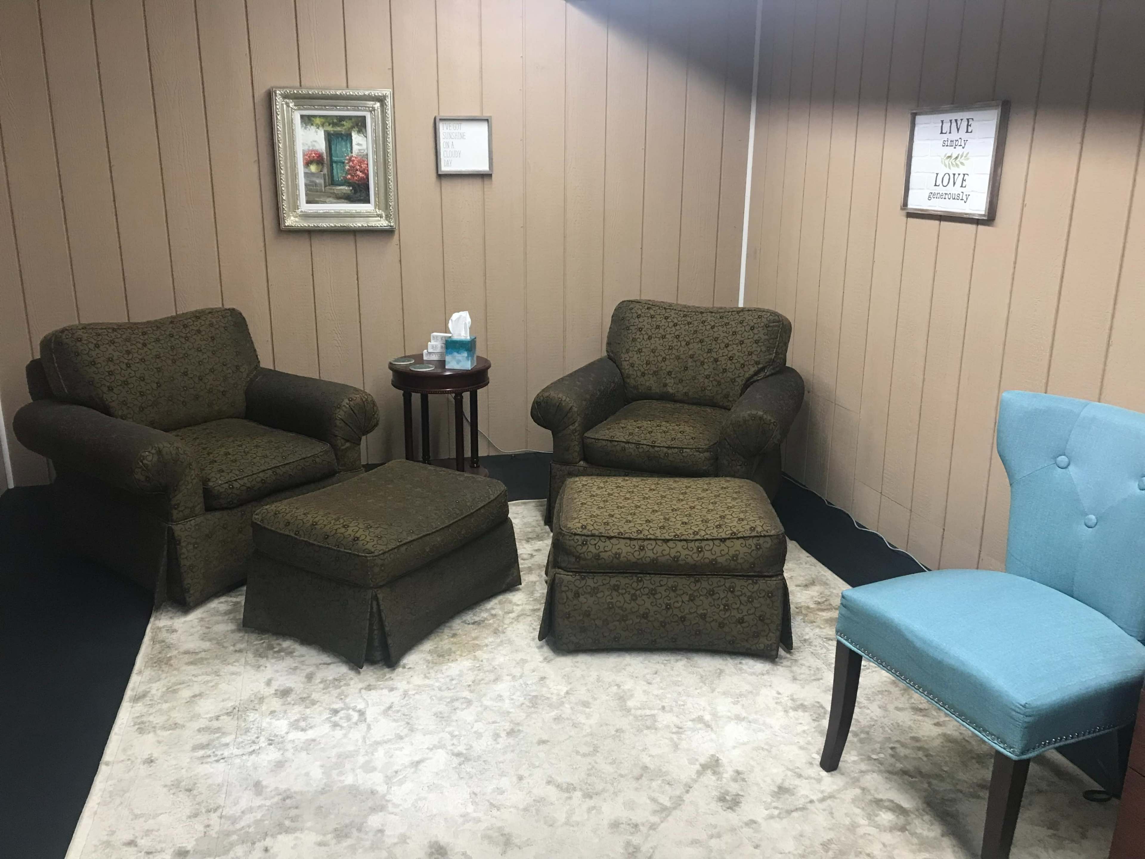 A seating area with two patterned armchairs, an ottoman, a small round table, a blue chair, and framed decorations on the walls.
