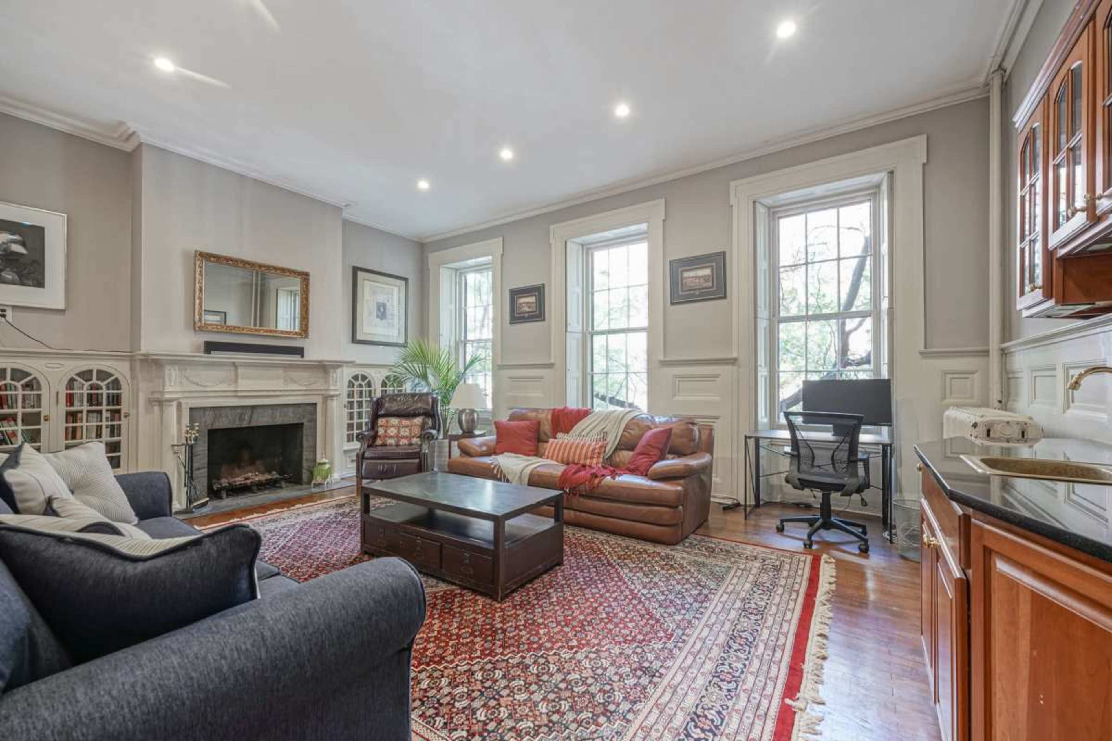 The image shows a living room with a fireplace, a coffee table, a sofa set, and a desk against the wall, all set on a large area rug.